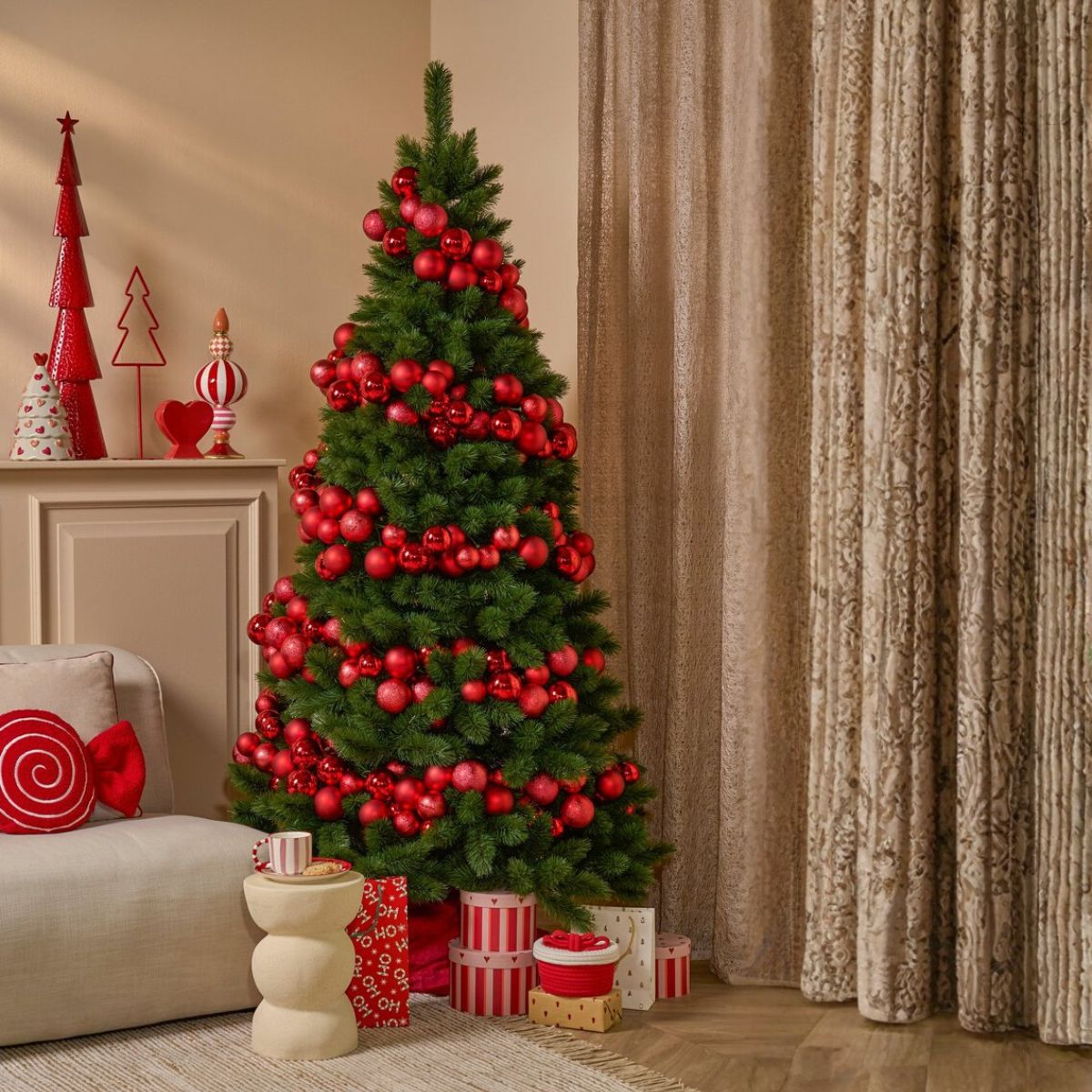 Een kerstboom versierd met rode ornamenten in een woonkamer naast een bank en cadeaus.
