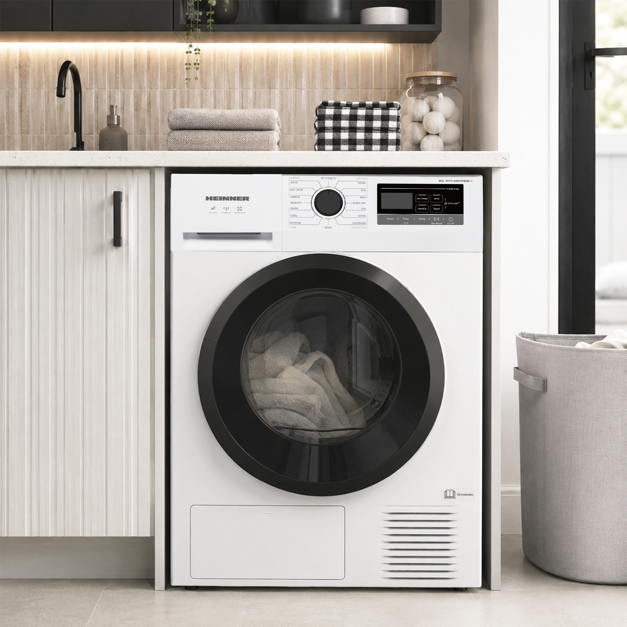 A white tumble dryer with a black door stands in a bright laundry room with shelves above.