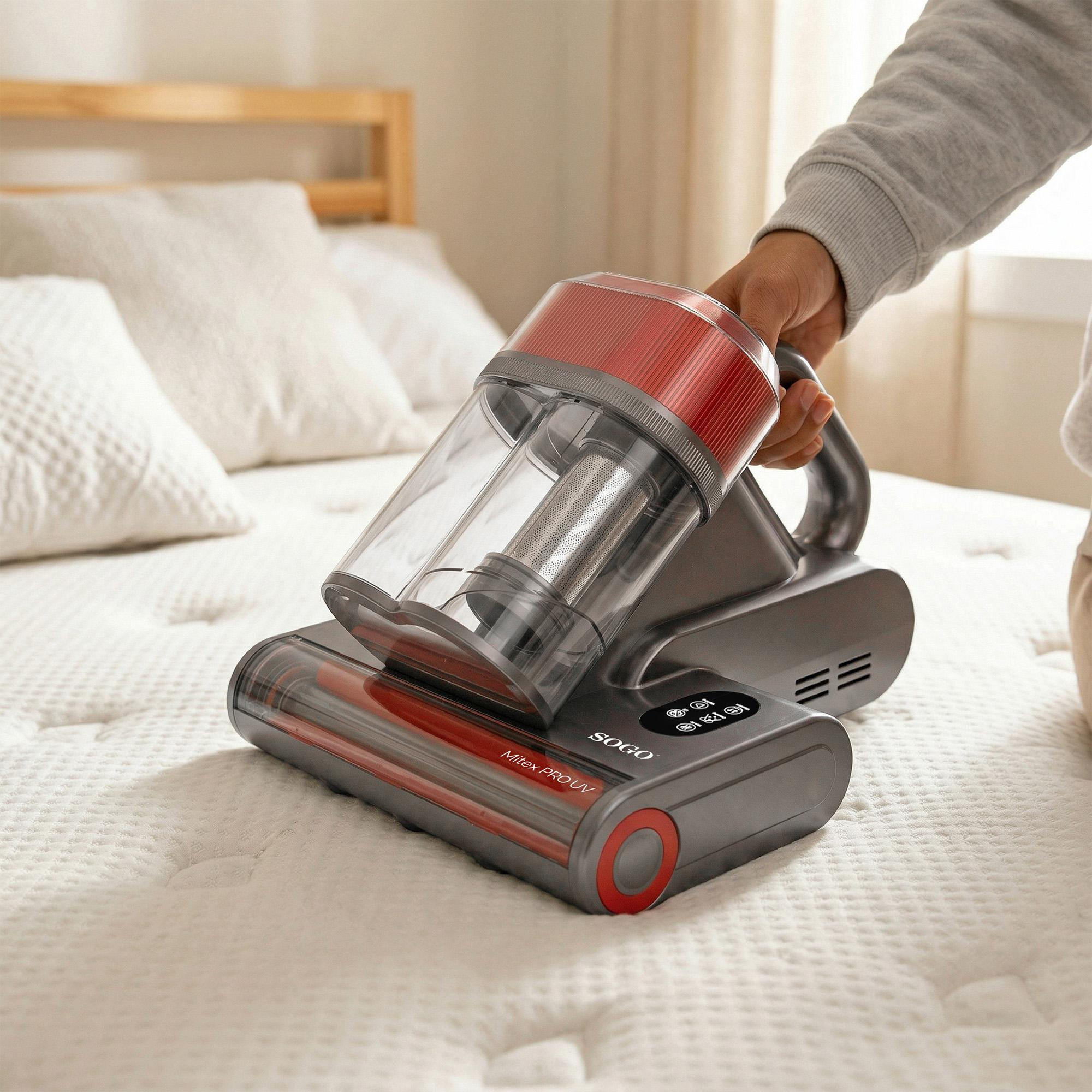 Una persona usa un aspirapolvere per materassi grigio e rosso a mano su un letto bianco.