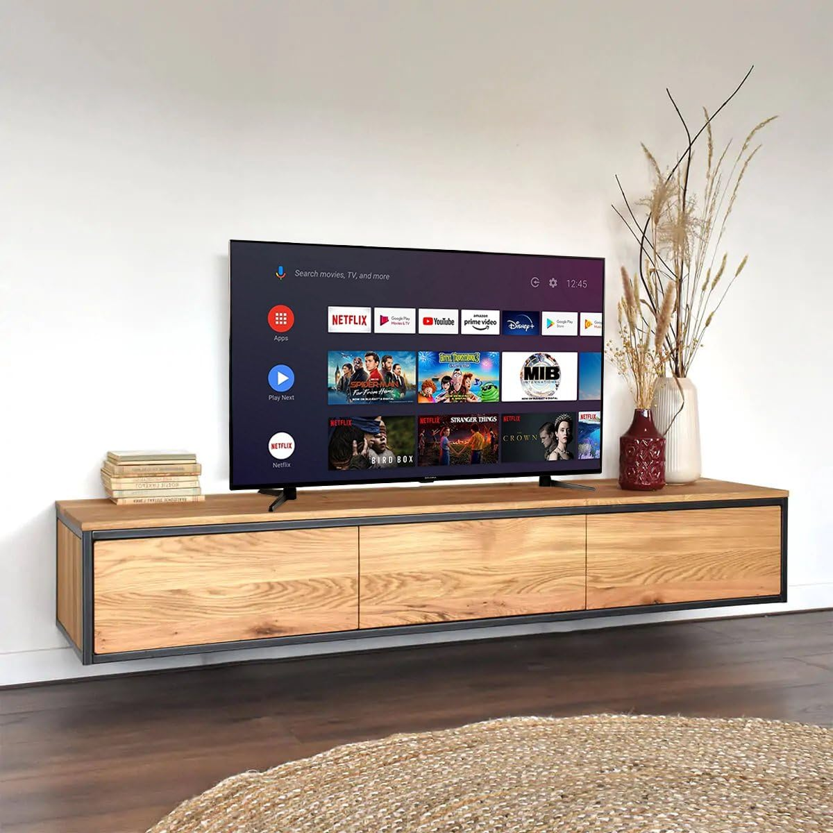 Un televisor con interfaz inteligente sobre un mueble de madera flotante, junto a un jarrón con flores secas.