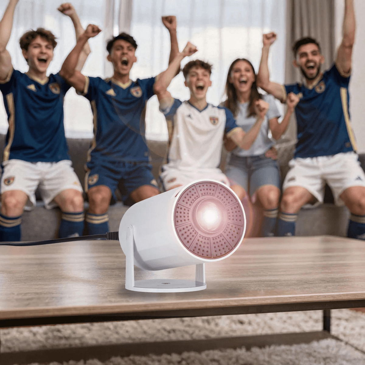 Ein weißer Projektor steht auf einem Holztisch, während jubelnde Fußballfans im Hintergrund sitzen.