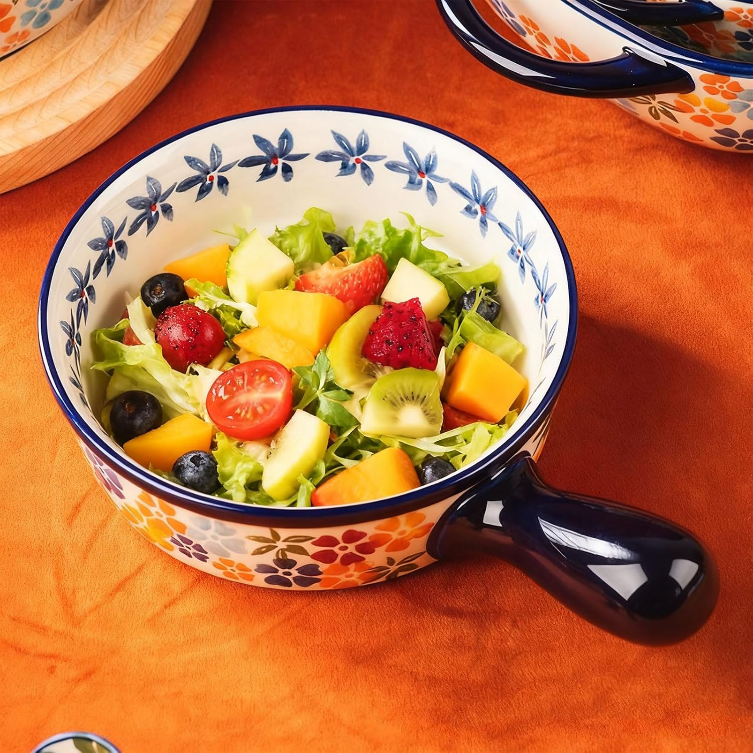 Eine Keramikschüssel mit Griff und Blumenmuster enthält einen bunten Obst- und Salatsalat auf einer orangefarbenen Fläche.