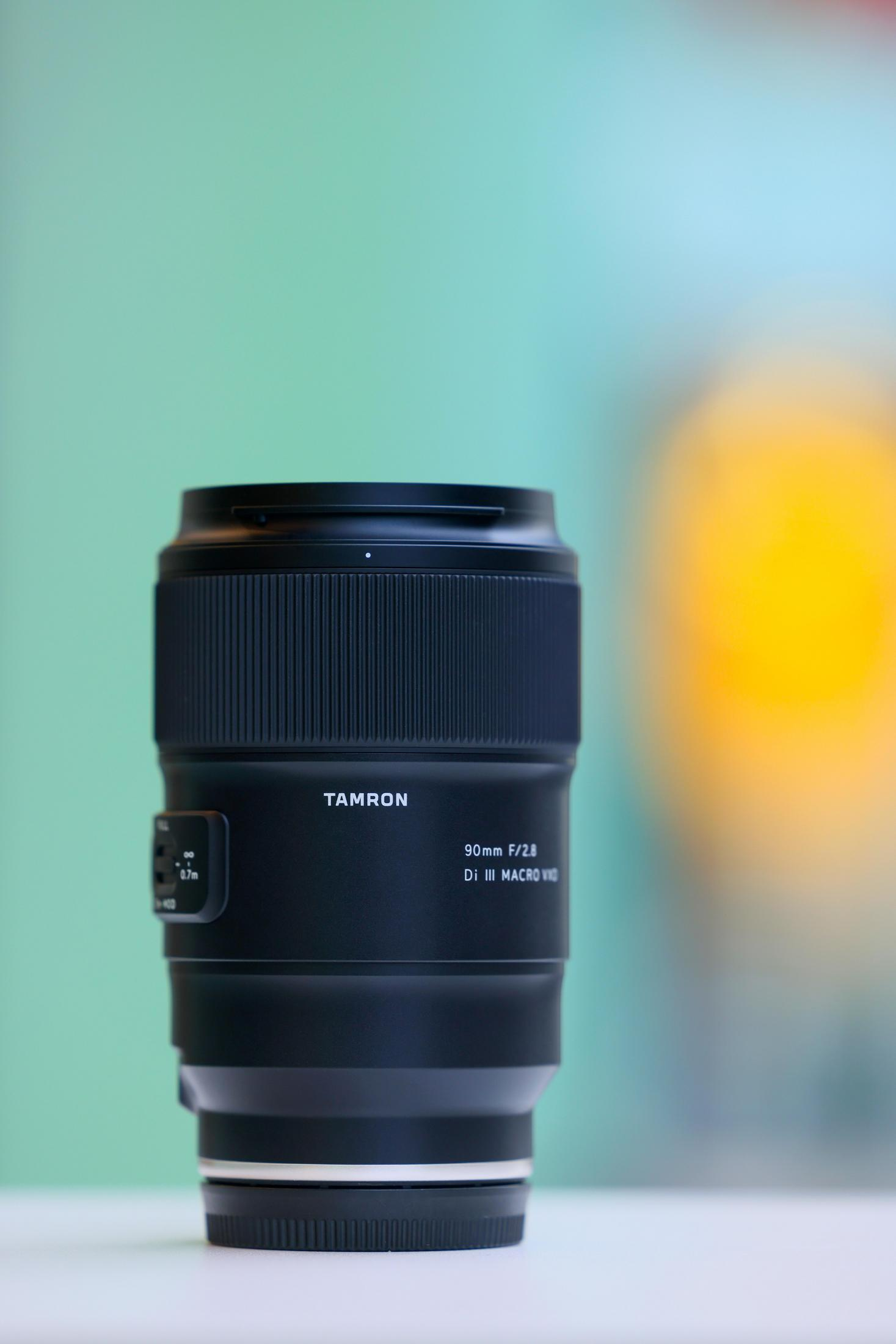 Ein schwarzes Tamron 90mm Makro-Kameraobjektiv steht aufrecht vor einem verschwommenen türkis-gelben Hintergrund.