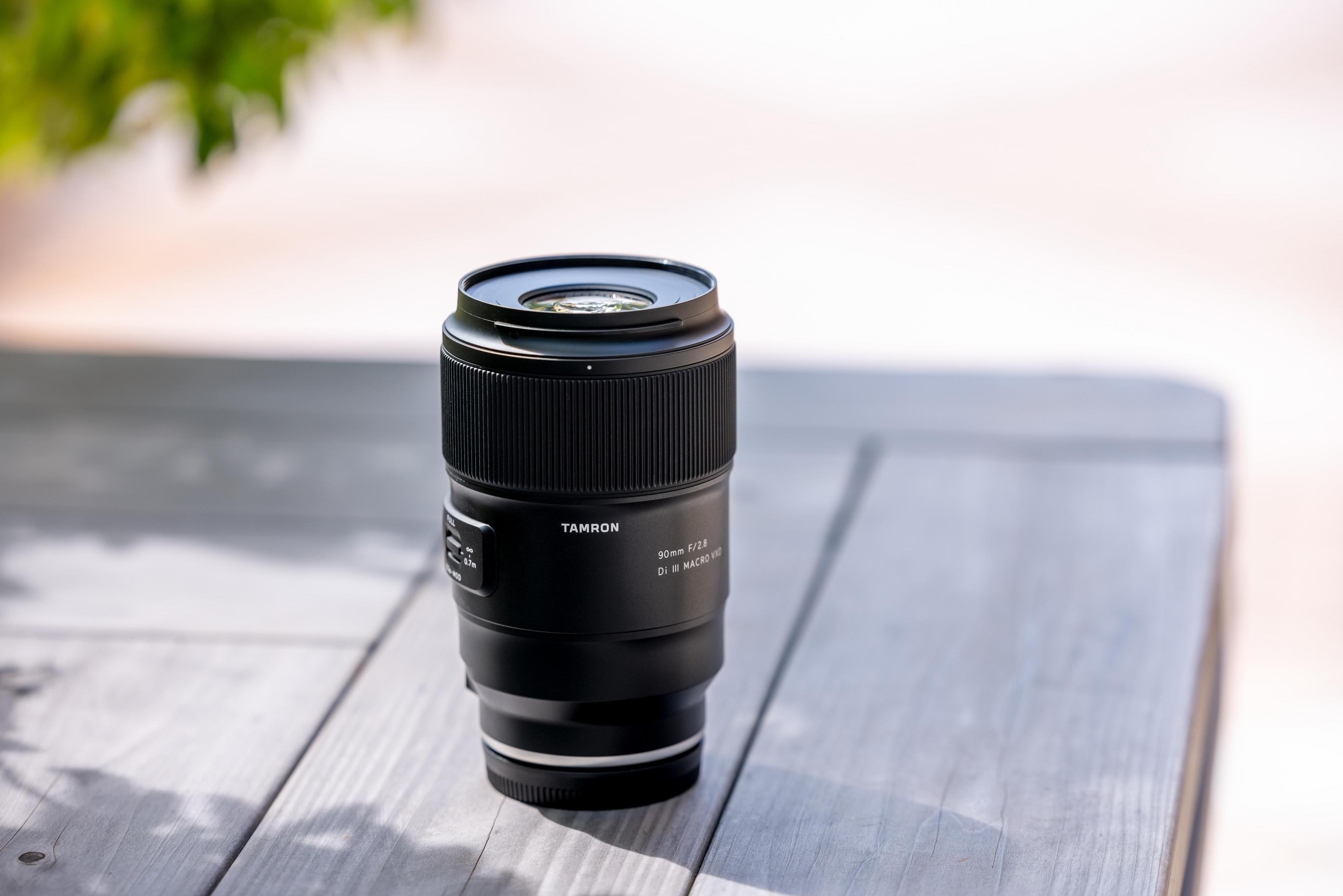 Ein schwarzes Tamron 90mm f/2.8 Macro Objektiv steht auf einem hölzernen Tisch im Freien.