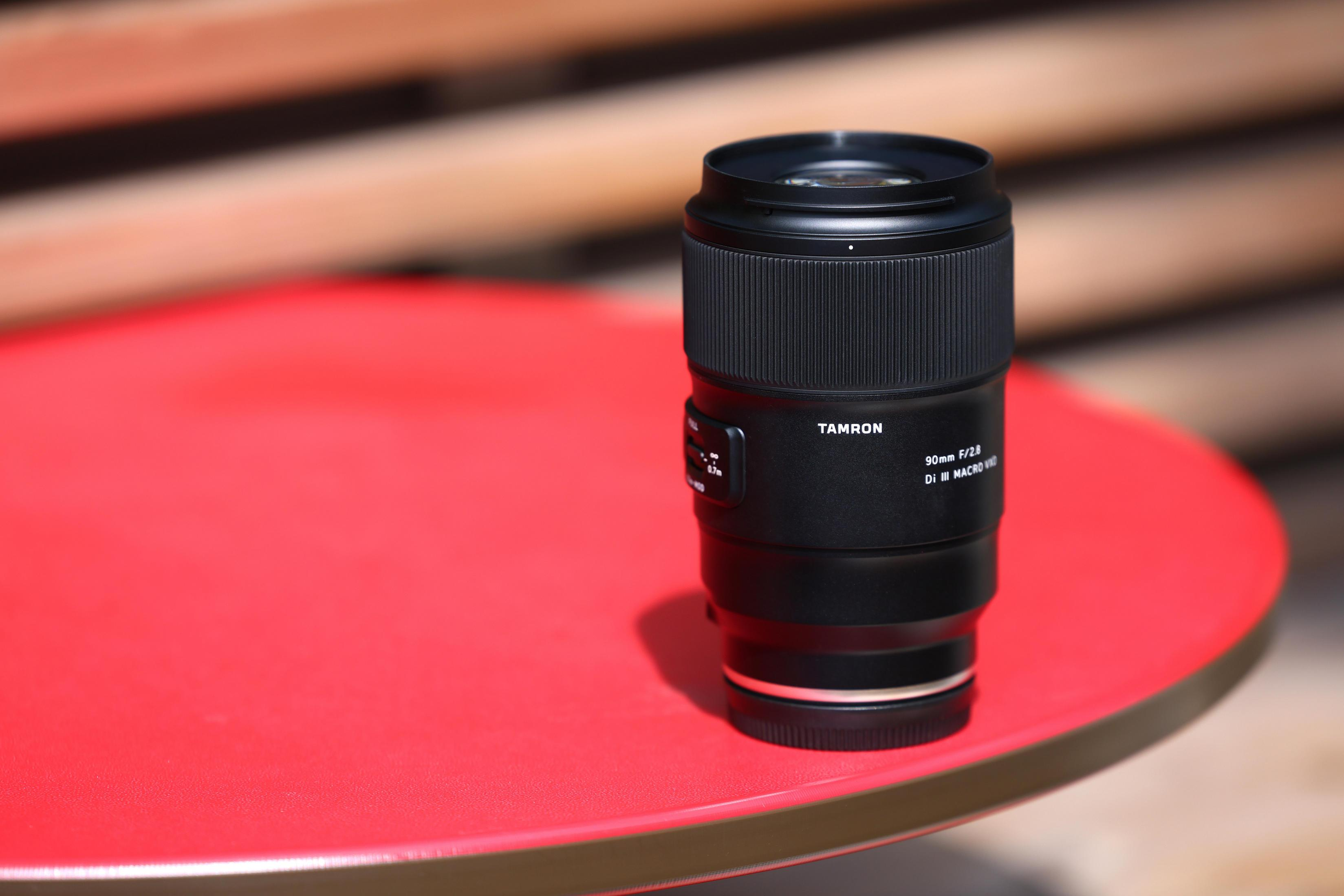Ein schwarzes Tamron 90mm Makro-Kameraobjektiv steht senkrecht auf einem runden roten Tisch im Freien.