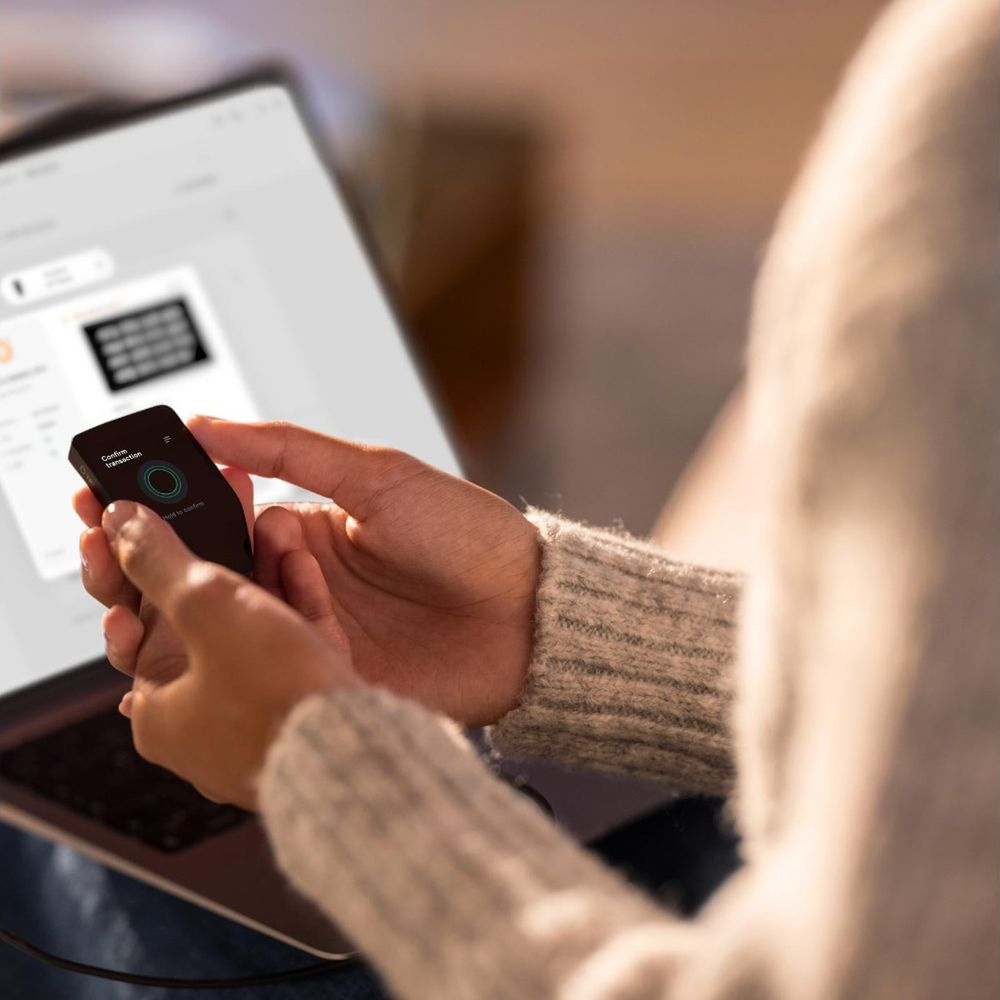 Person hält ein kleines schwarzes Gerät vor einem Laptop, das eine Transaktion bestätigt.
