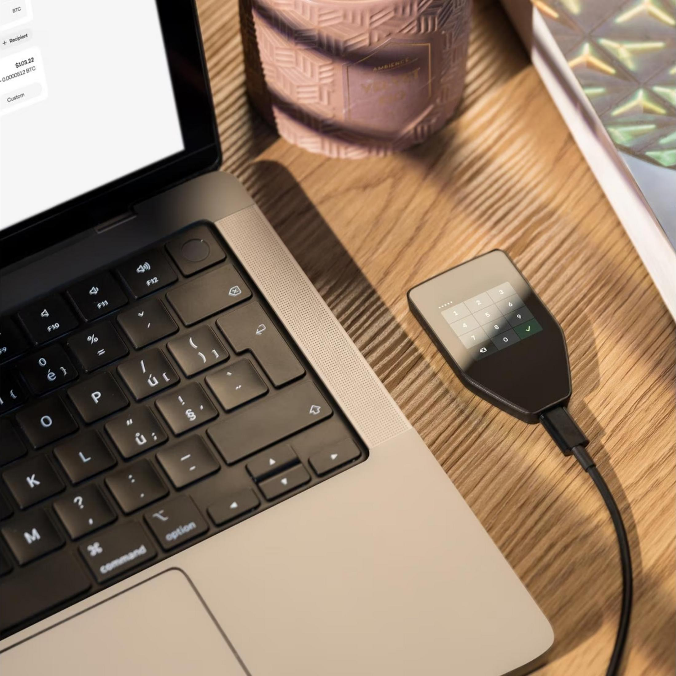 Ein Laptop, eine Kerze und ein Krypto-Hardware-Wallet mit Tastatur liegen auf einem Holztisch.