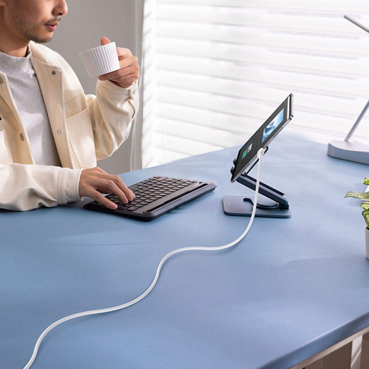 Un uomo beve caffè a una scrivania blu, digitando su una tastiera vicino a un tablet su supporto.