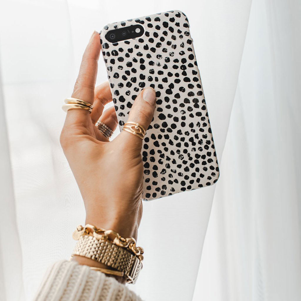 Una mano con anelli e bracciali d'oro tiene uno smartphone bianco con pois neri davanti a una tenda.