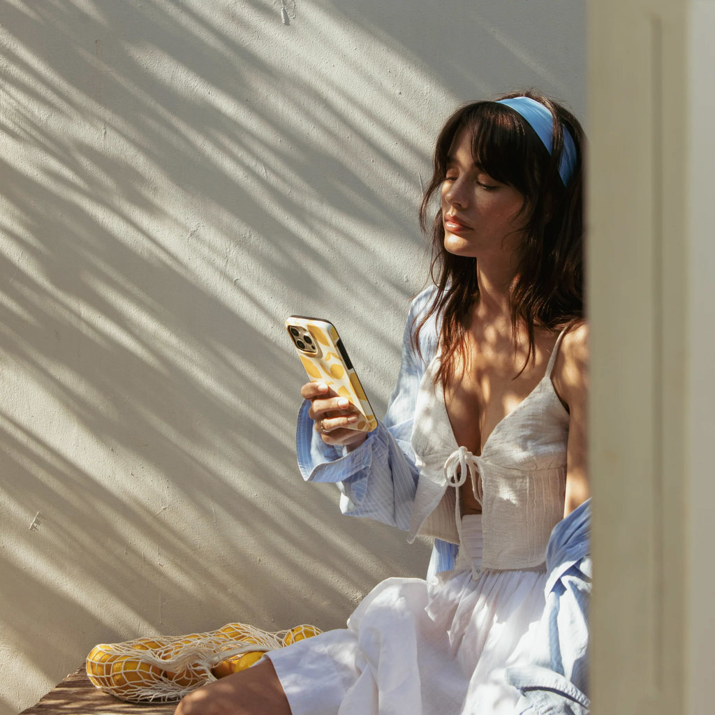 Une femme en blanc avec un headband bleu regarde son téléphone près d'un mur aux ombres d'arbres.