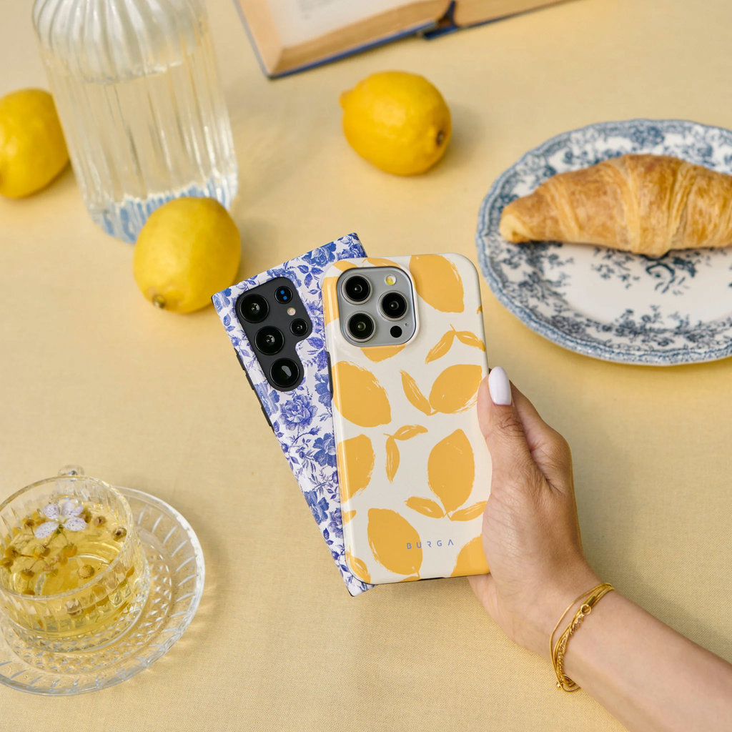 Une main tient deux coques de téléphone à motifs de citrons et de fleurs bleues sur une table jaune avec des citrons, du thé et un croissant.