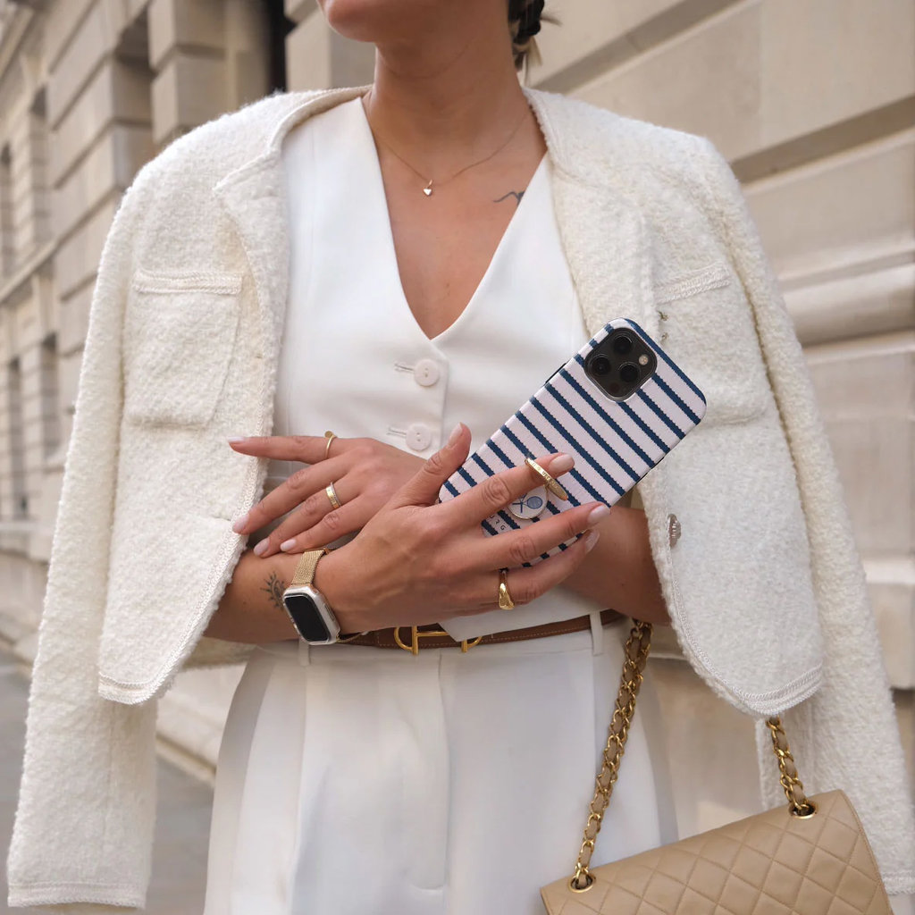 Een vrouw in witte kleding houdt een blauw-wit gestreepte smartphone en een beige handtas vast.