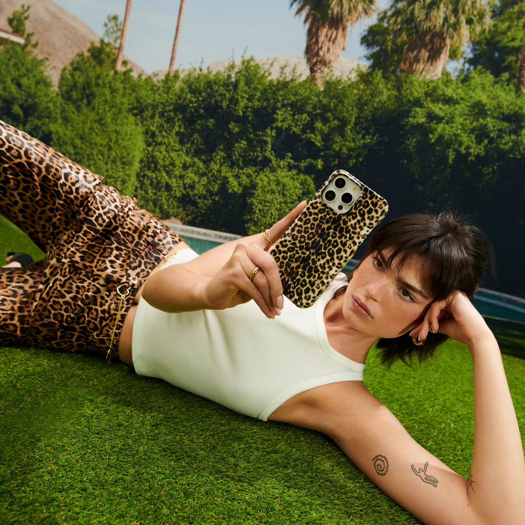 Una donna con pantaloni leopardati e top bianco sdraiata sull'erba tiene in mano un telefono coordinato.