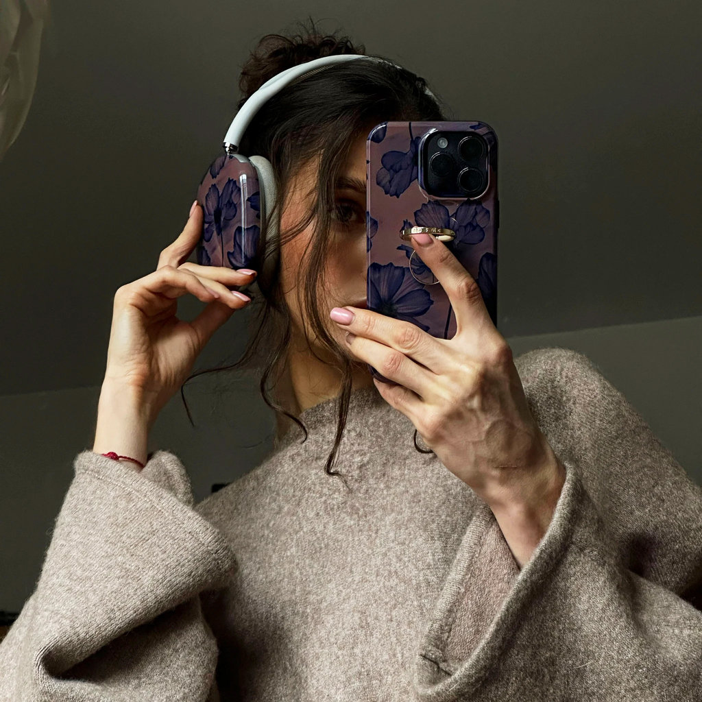 Une femme en pull beige tient un téléphone fleuri devant son visage et porte des écouteurs assortis.
