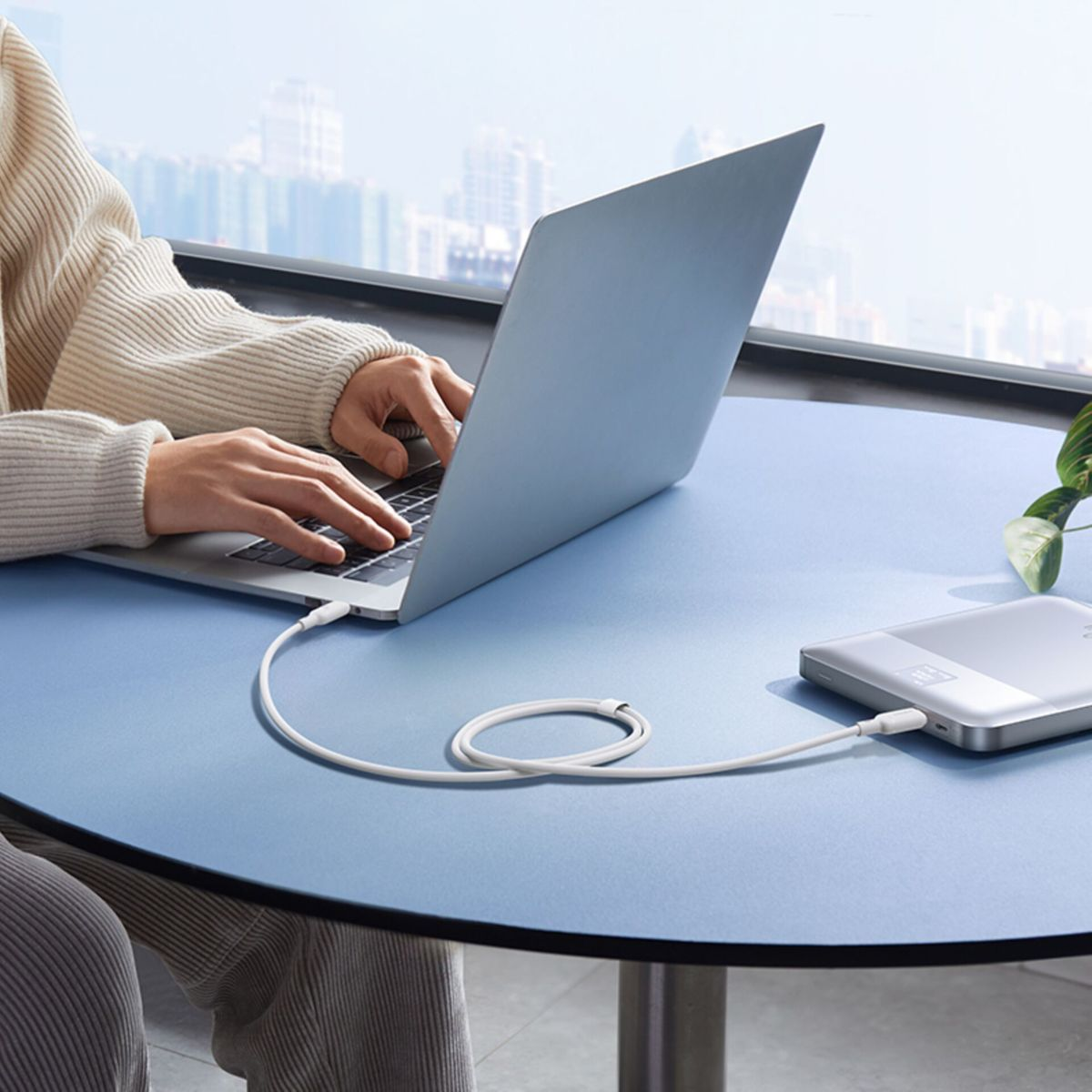 Persona con maglione bianco digita su laptop collegato con cavo bianco a power bank su tavolo blu.