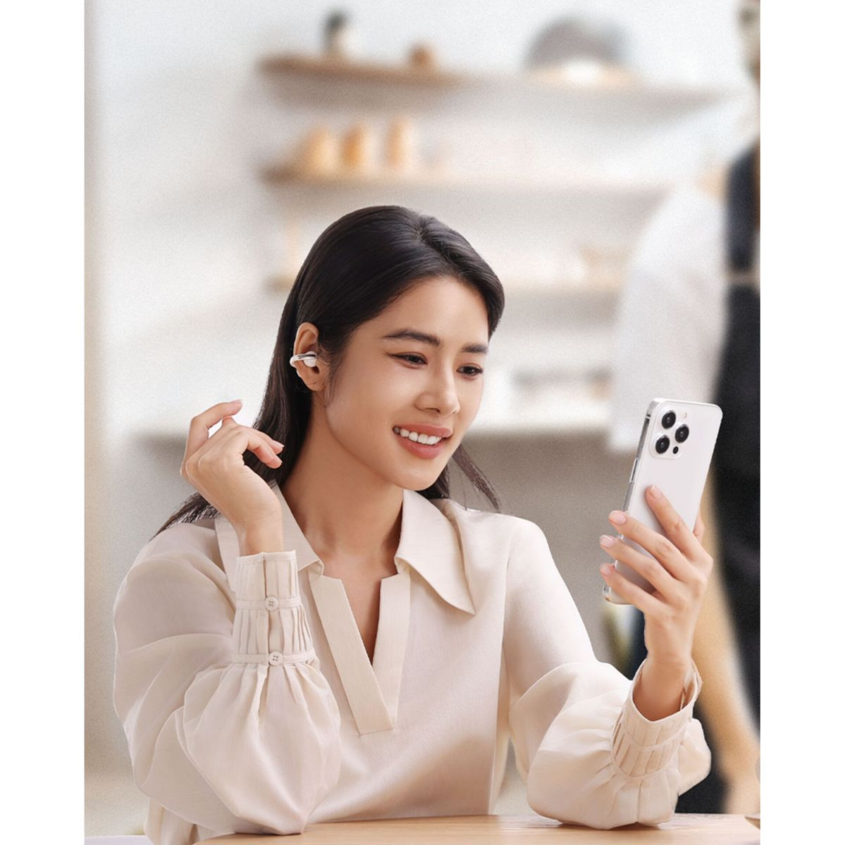 Una donna sorridente con auricolari bianchi guarda il suo smartphone al tavolo.