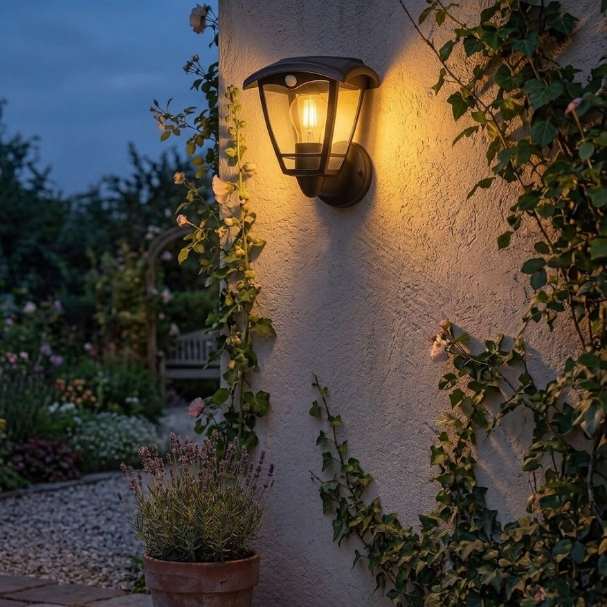Beleuchtete Wandleuchte im Garten an einer weißen Wand mit grünen Pflanzen im Hintergrund bei Dämmerung.