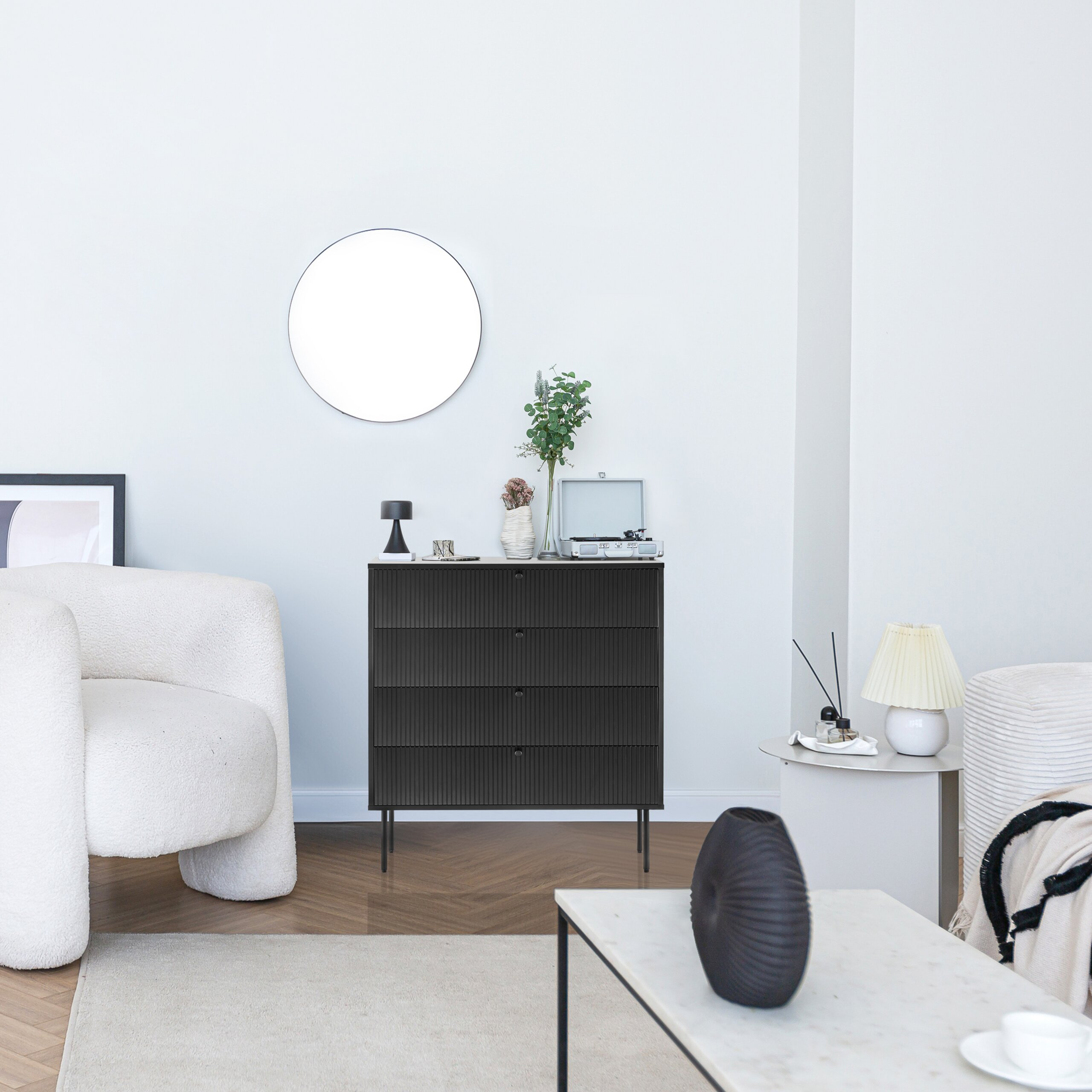Moderne Einrichtung mit weißem Sessel, schwarzer Kommode und runden Spiegel an weißer Wand.