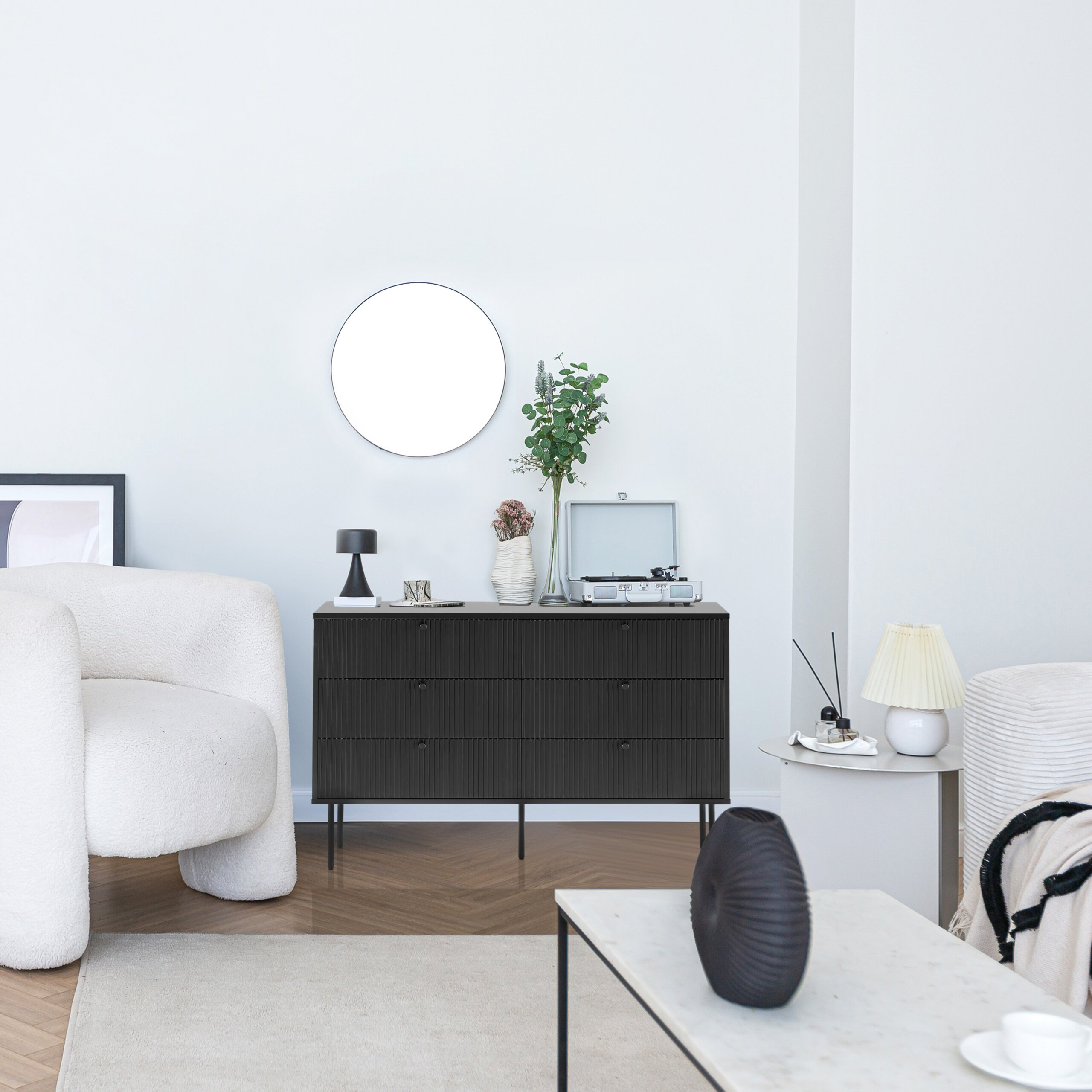 Ein minimalistisches Wohnzimmer mit einer dunklen Kommode, einem runden Spiegel und cremefarbenen Sesseln.