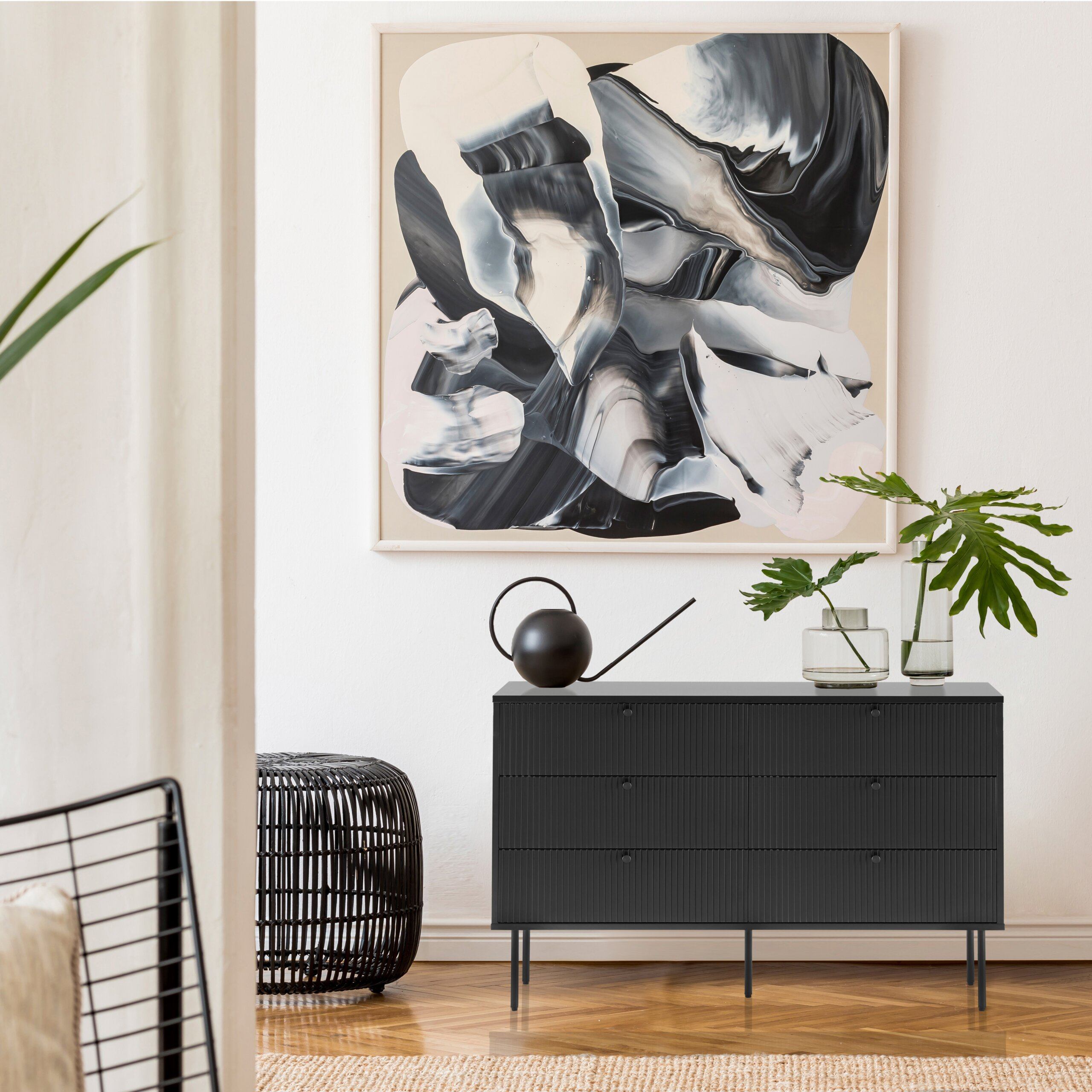 Eine schwarze Kommode mit Gießkanne, Glasvasen mit Pflanzen und einem großen abstrakten Gemälde an der weißen Wand.