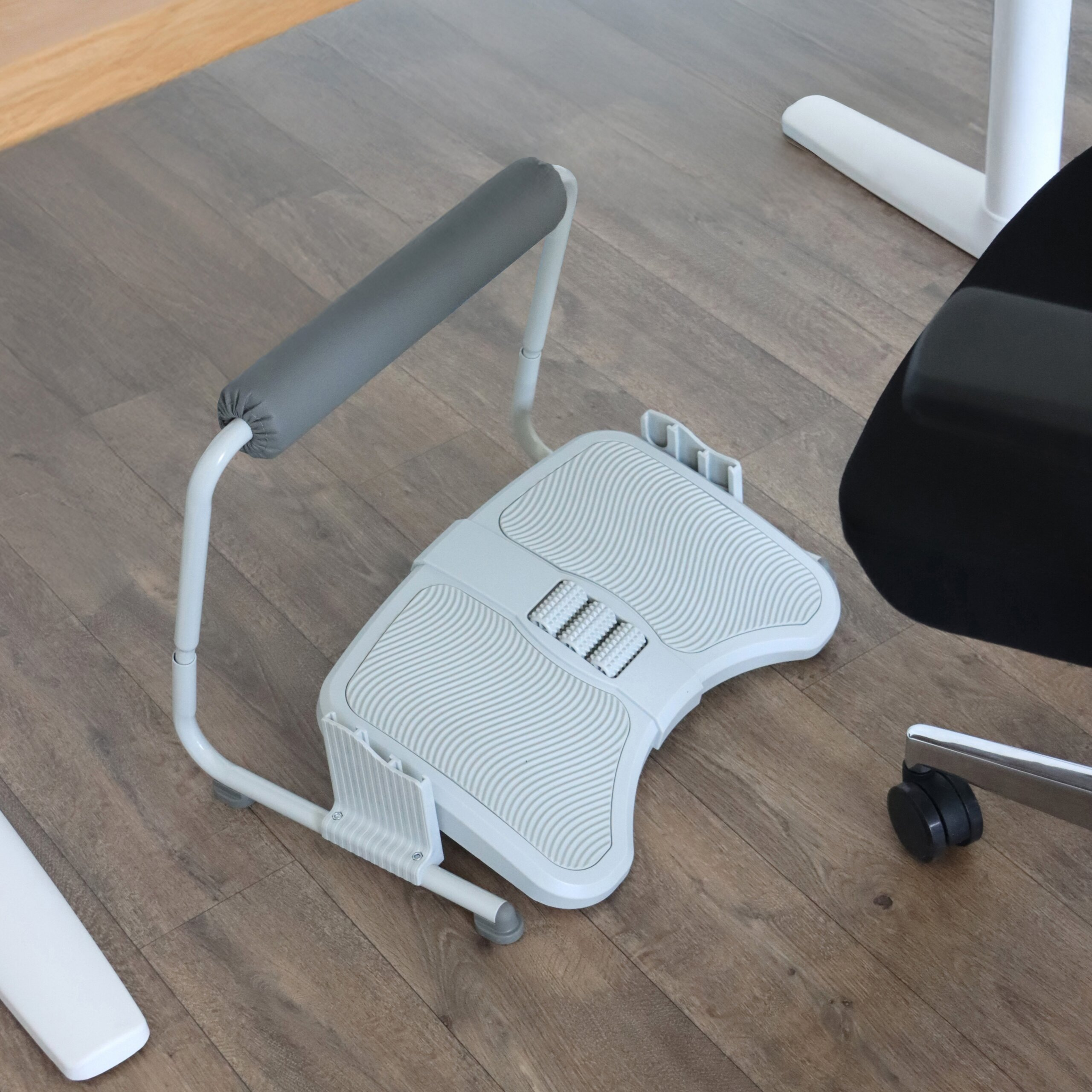 Eine weiße ergonomische Fußstütze mit Massagegängern und grauer Wadenpolsterung auf Holzboden.