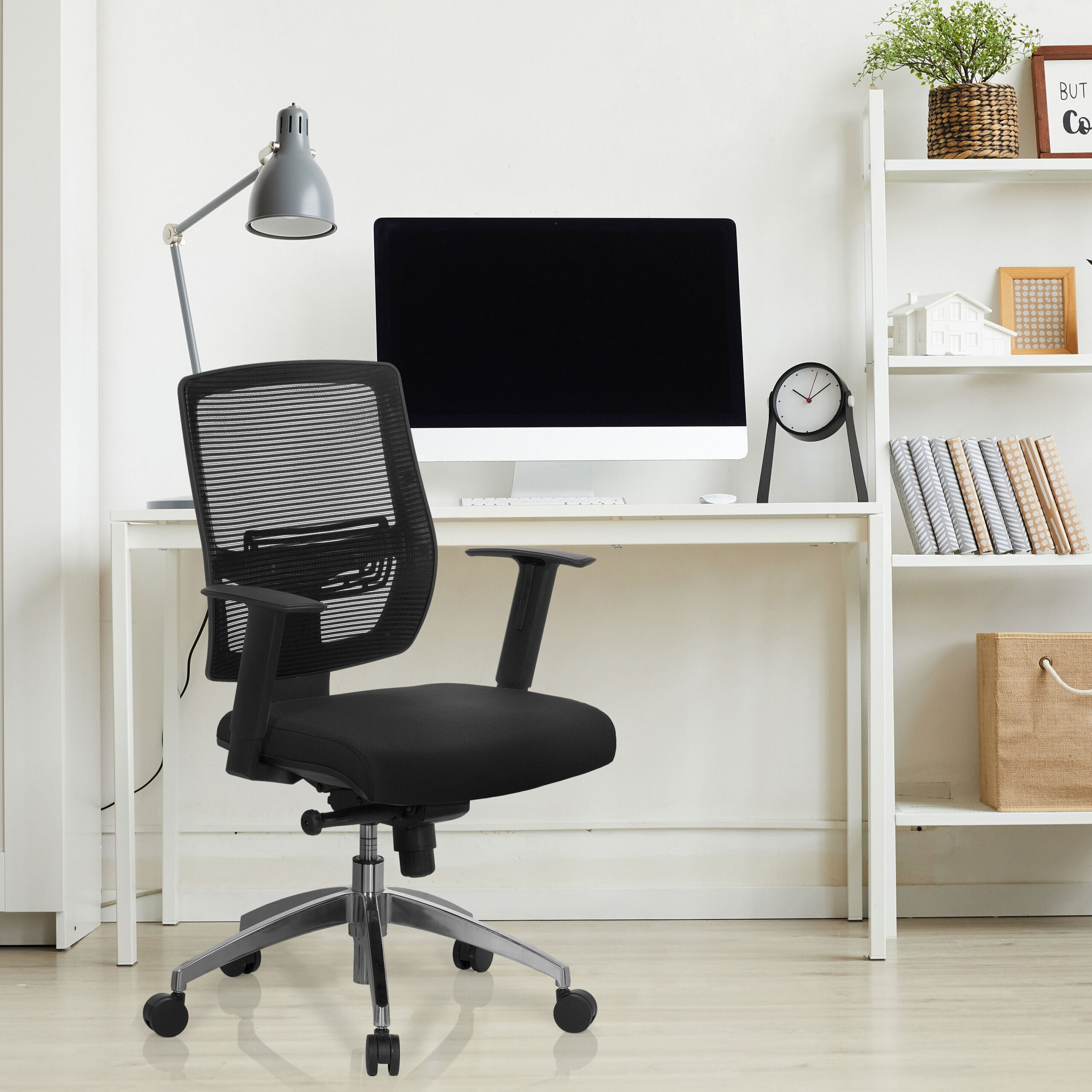 Ein schwarzer Netz-Bürostuhl steht vor einem weißen Schreibtisch mit Computer und Lampe. Rechts ein weißes Regal.