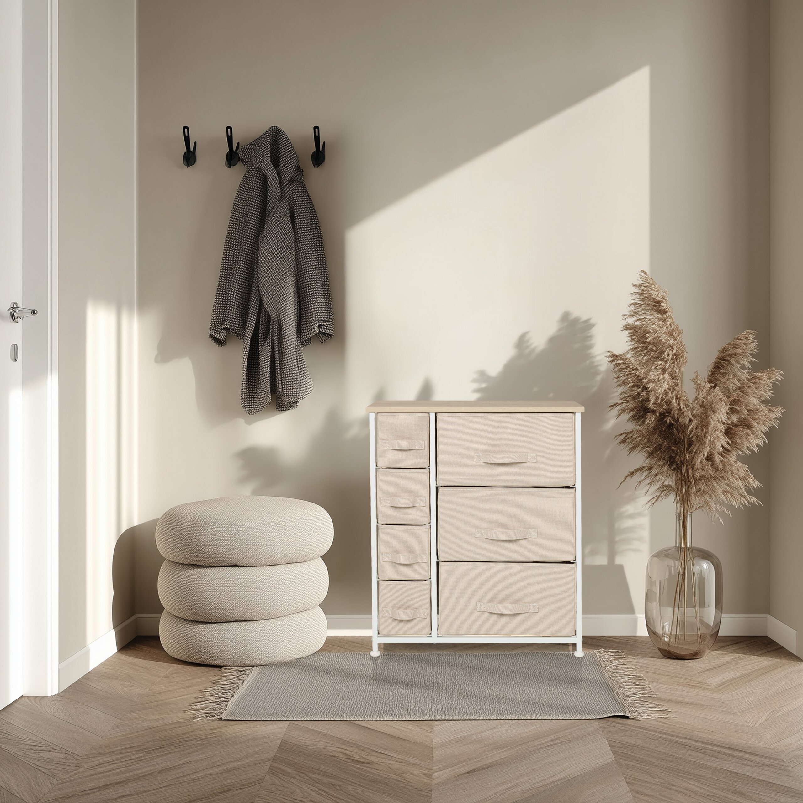 Ein beige Flur mit Mantel an Wandhaken, Stoffkommode, Sitzpuff und getrocknetem Gras in Glasvase.