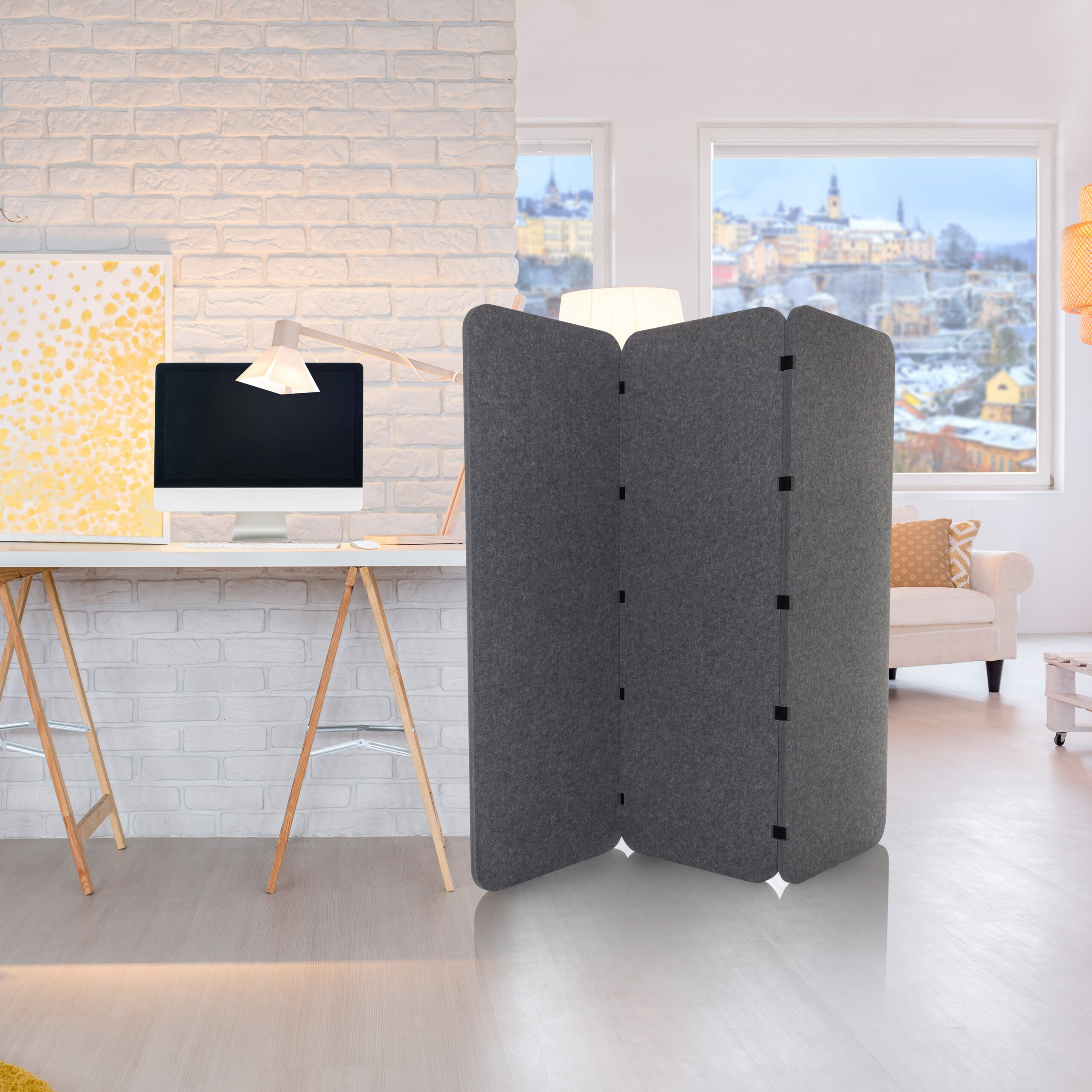 Ein grauer Raumteiler steht in einem hellen Zimmer vor einem Schreibtisch mit Computer und einer Ziegelwand.