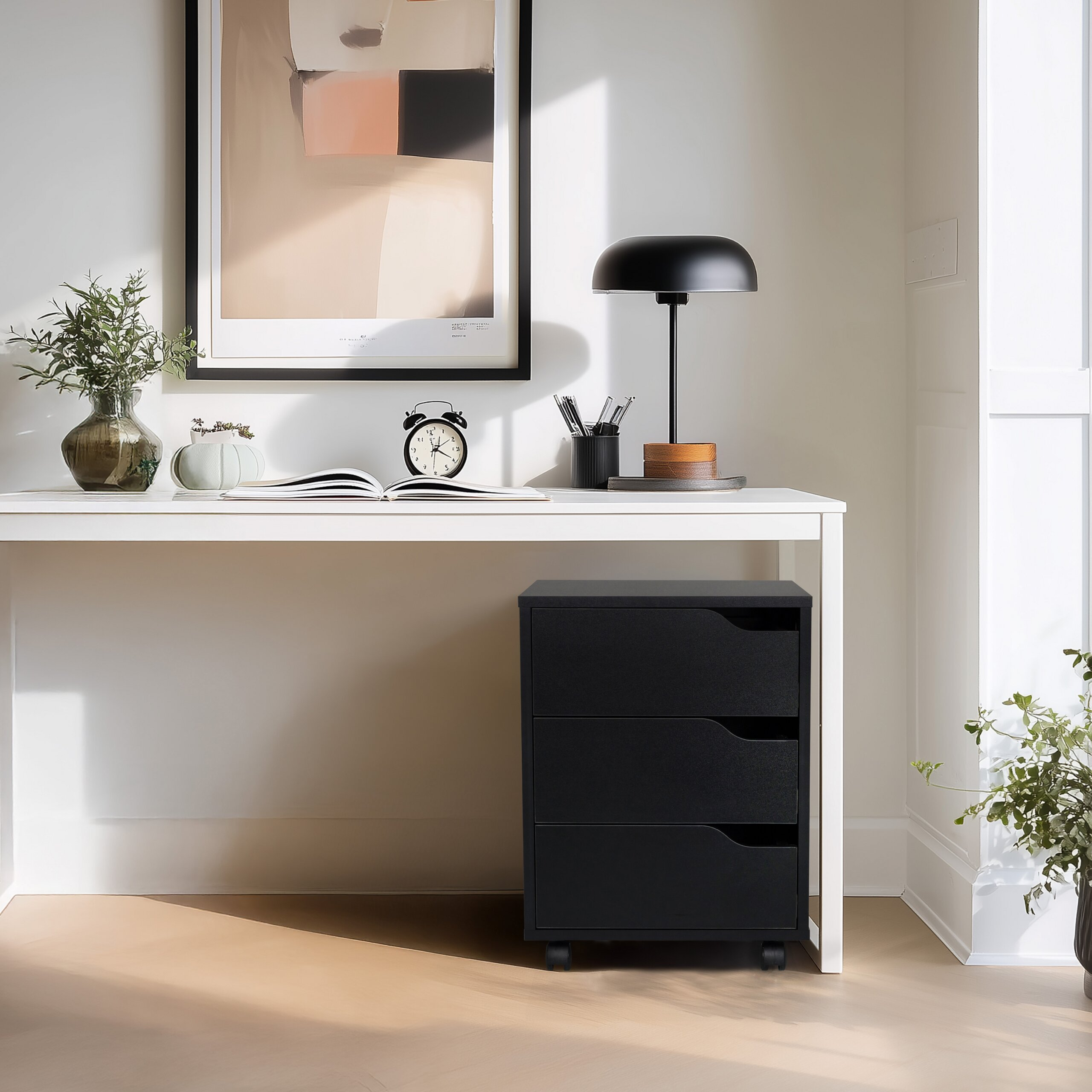 Ein weißer Schreibtisch mit schwarzem Rollcontainer, Lampe, Uhr und gerahmtem Bild an der Wand.