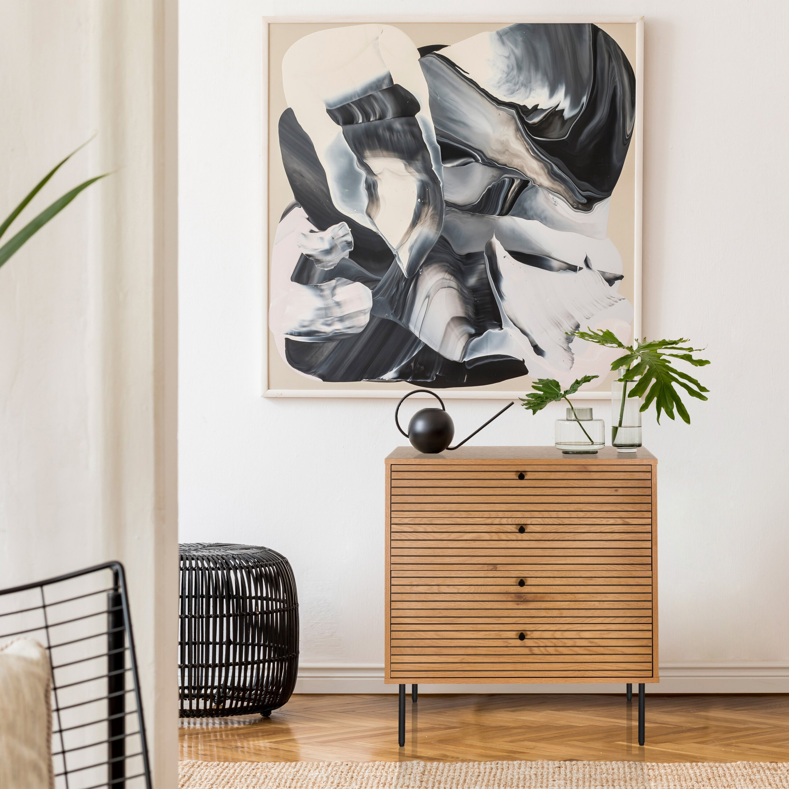 Eine Holzkommode mit Pflanzen und einer Gießkanne vor einem großen abstrakten Bild an einer weißen Wand.