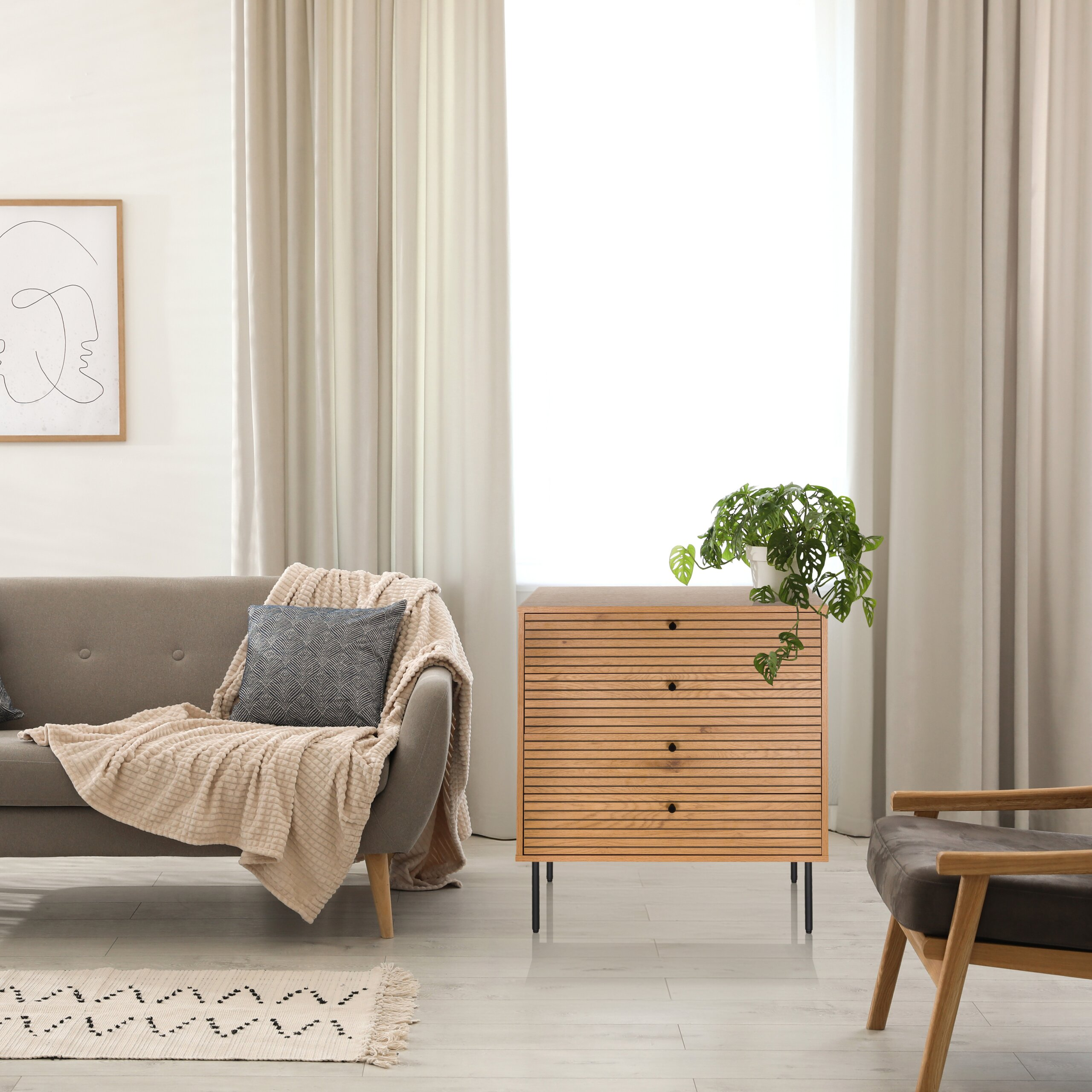 Ein helles Wohnzimmer mit grauer Couch, Holzkommode, Zimmerpflanze, Wandkunst und großen Vorhängen vor Fenstern.