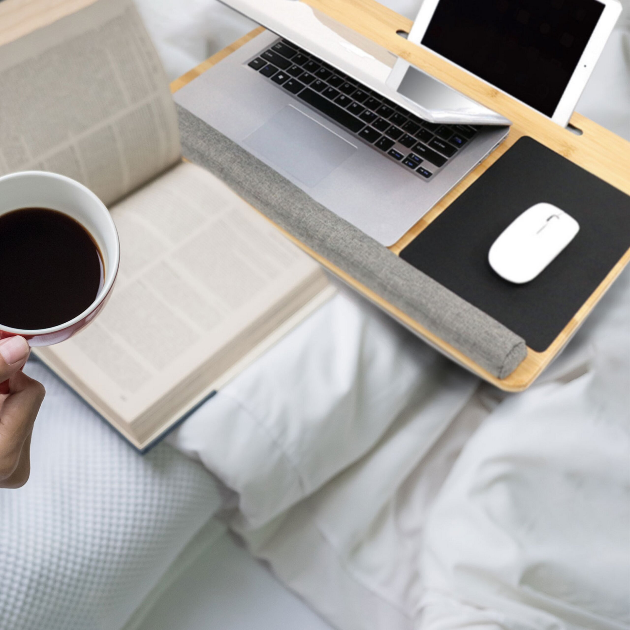 Holztisch auf dem Bett mit Laptop, Tablet, Maus und offener Buch, Person hält eine Kaffeetasse.