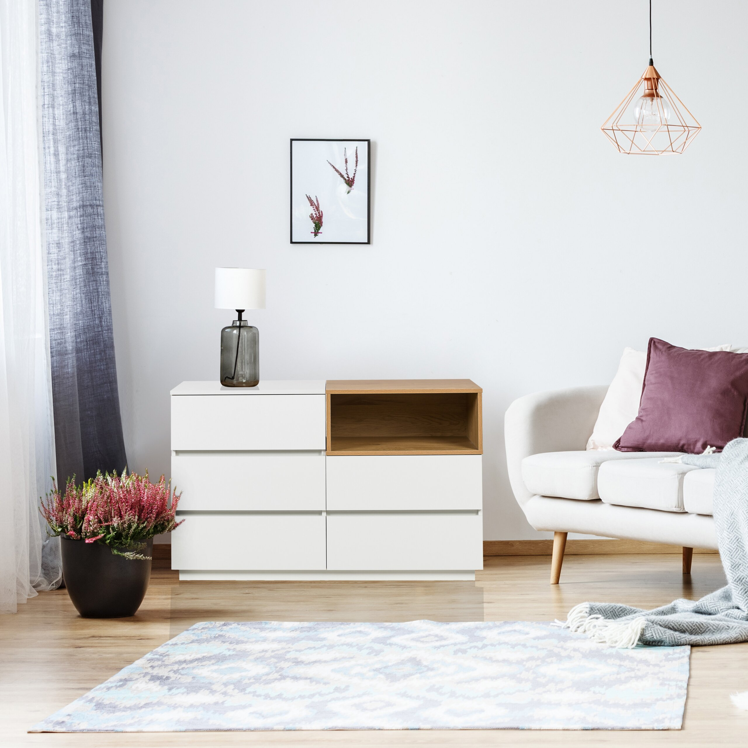 Ein weißer Schrank, ein Sofa mit lilafarbenem Kissen und eine Topfpflanze stehen in einem hellen Wohnzimmer.
