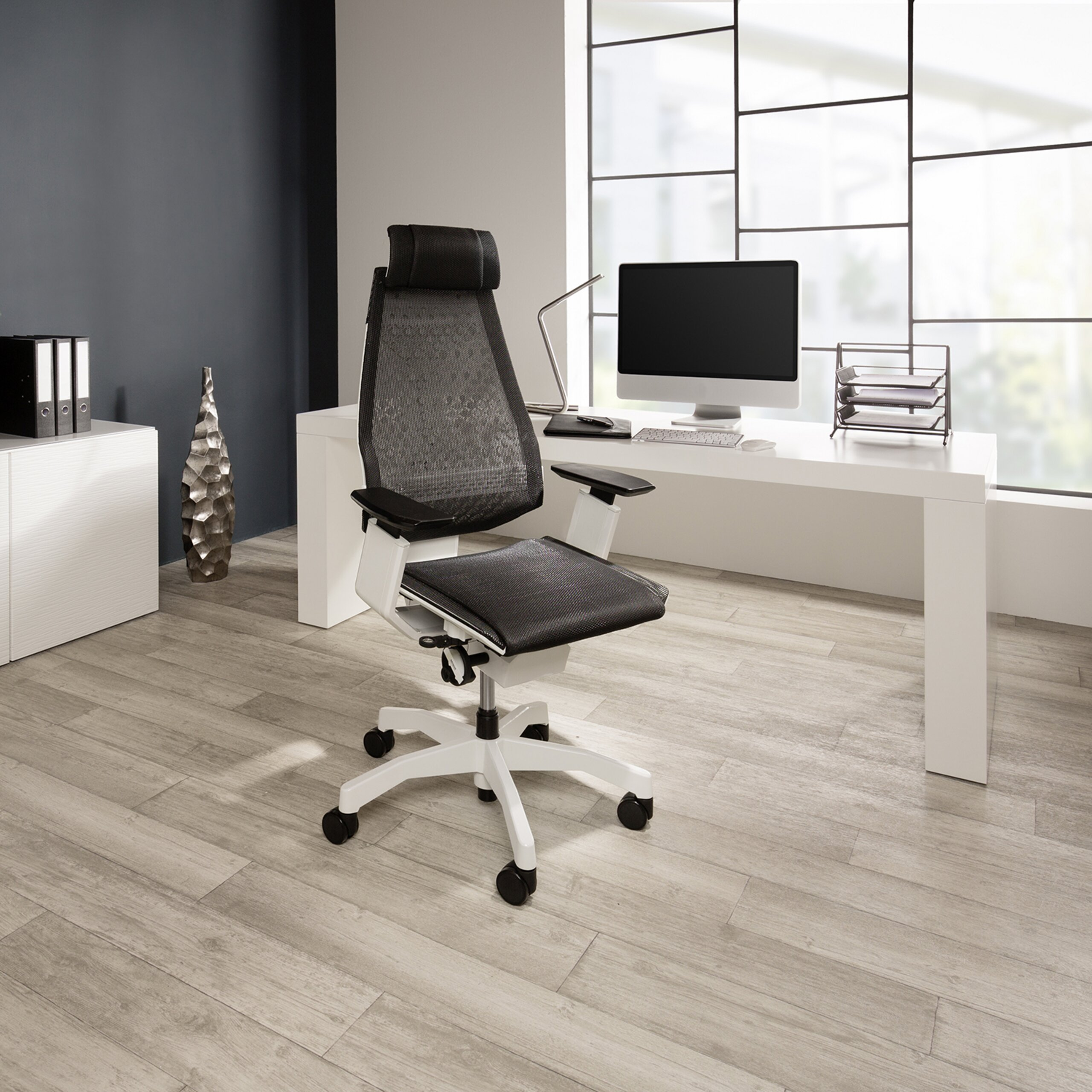 Ein schwarz-weißer ergonomischer Buerostuhl steht vor einem weißen Schreibtisch mit Computer und grossem Fenster.