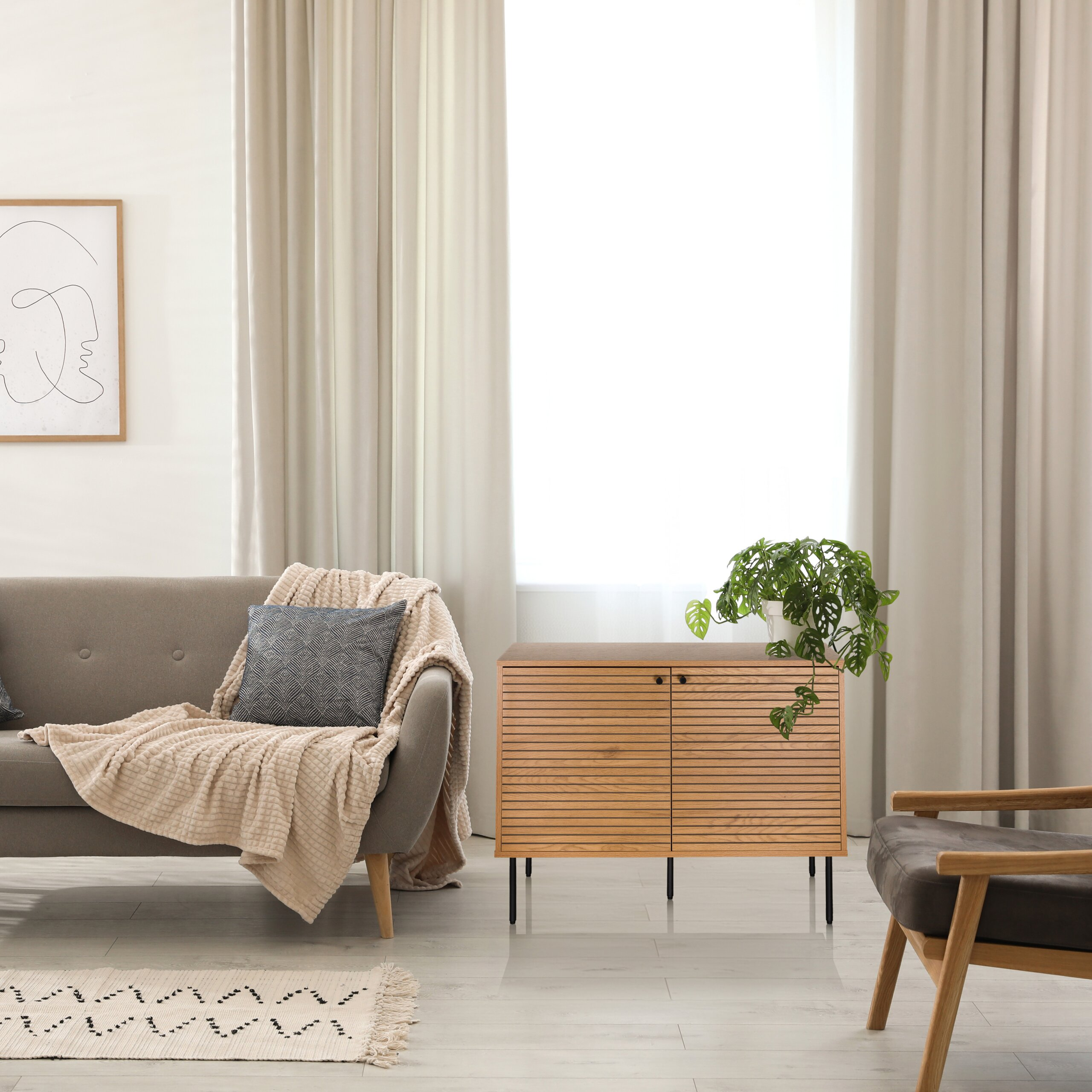 Ein gemütliches Wohnzimmer mit grauer Couch, beigem Plaid, Holzkommode mit Pflanze und großen Fenstern mit Vorhängen.