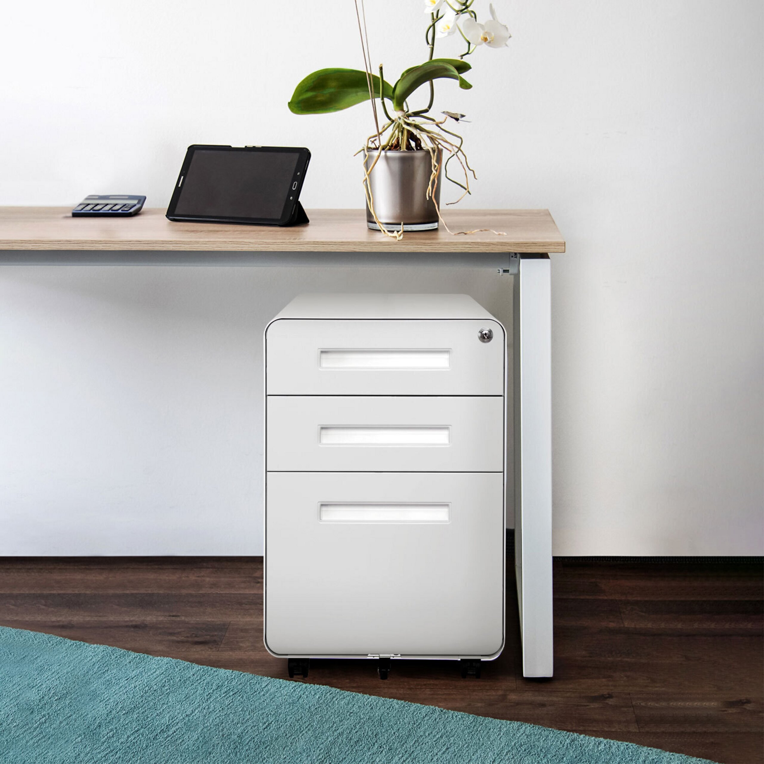 Ein weißer Rollcontainer steht unter einem Holztisch mit Tablet, Rechner und Orchidee.