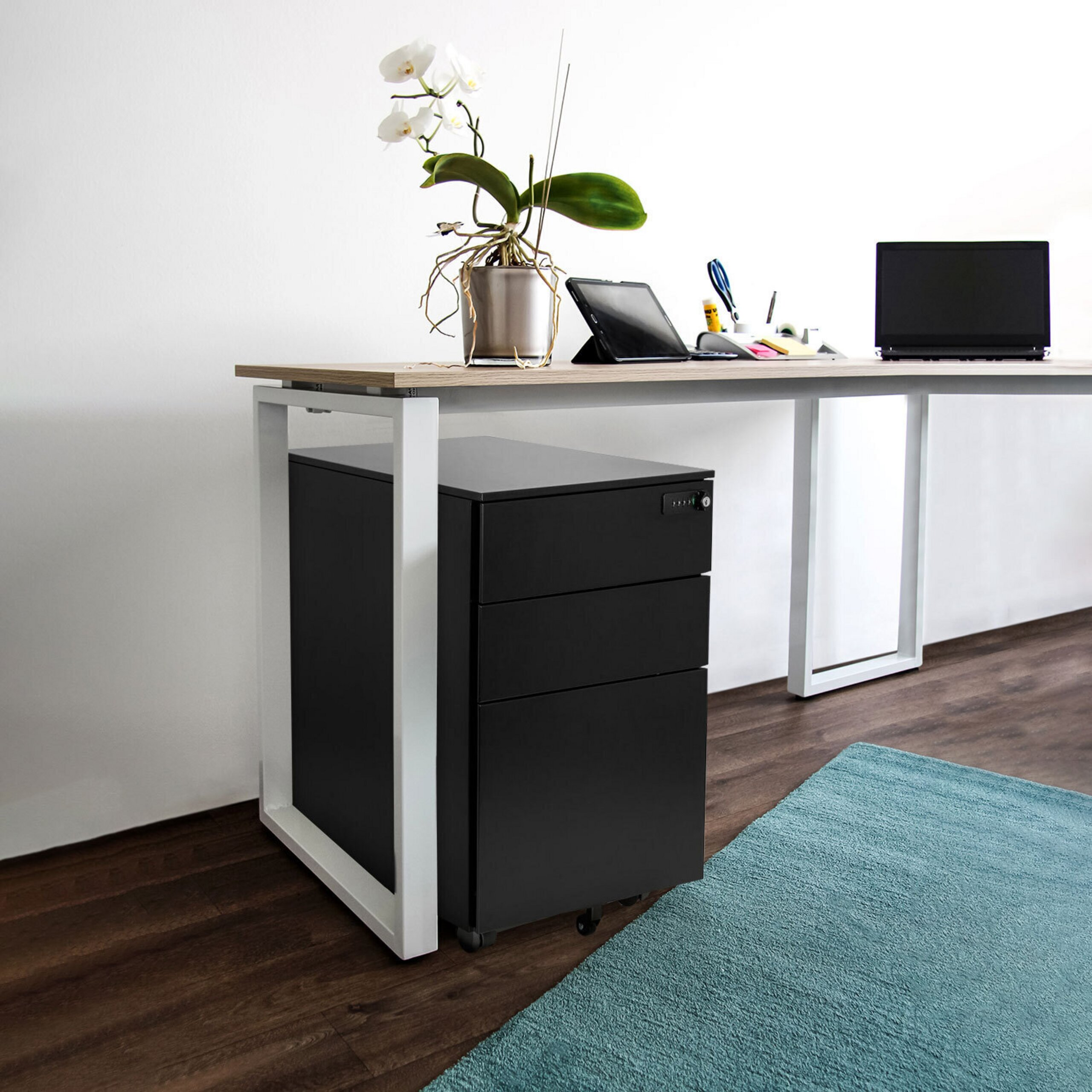 Ein moderner Schreibtisch mit weißem Gestell, schwarzem Rollcontainer, Laptop, Tablet und einer weißen Orchidee.