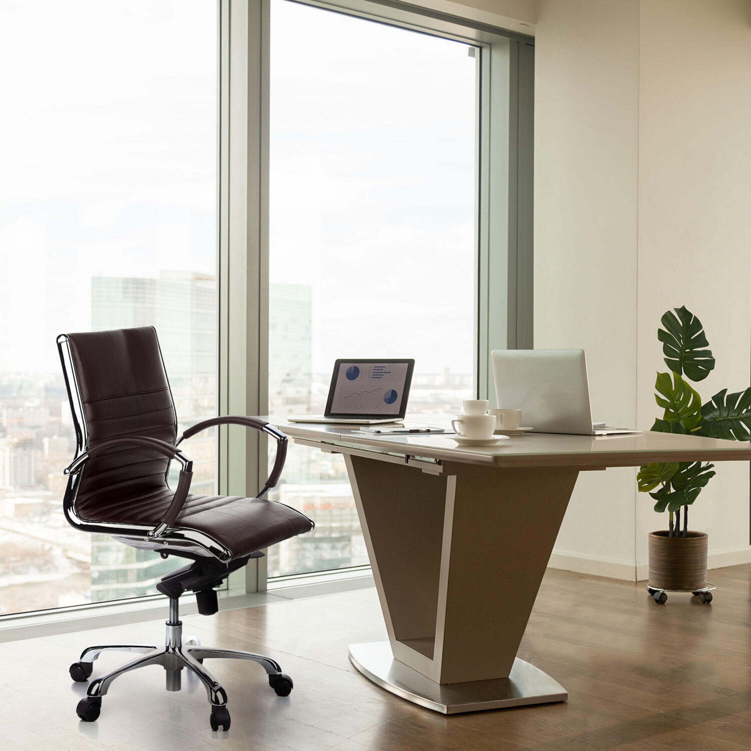 Moderne Bueroumgebung mit einem dunklen Ledersessel, einem Tisch mit Laptops und einer Topfpflanze vor grossem Fenster.