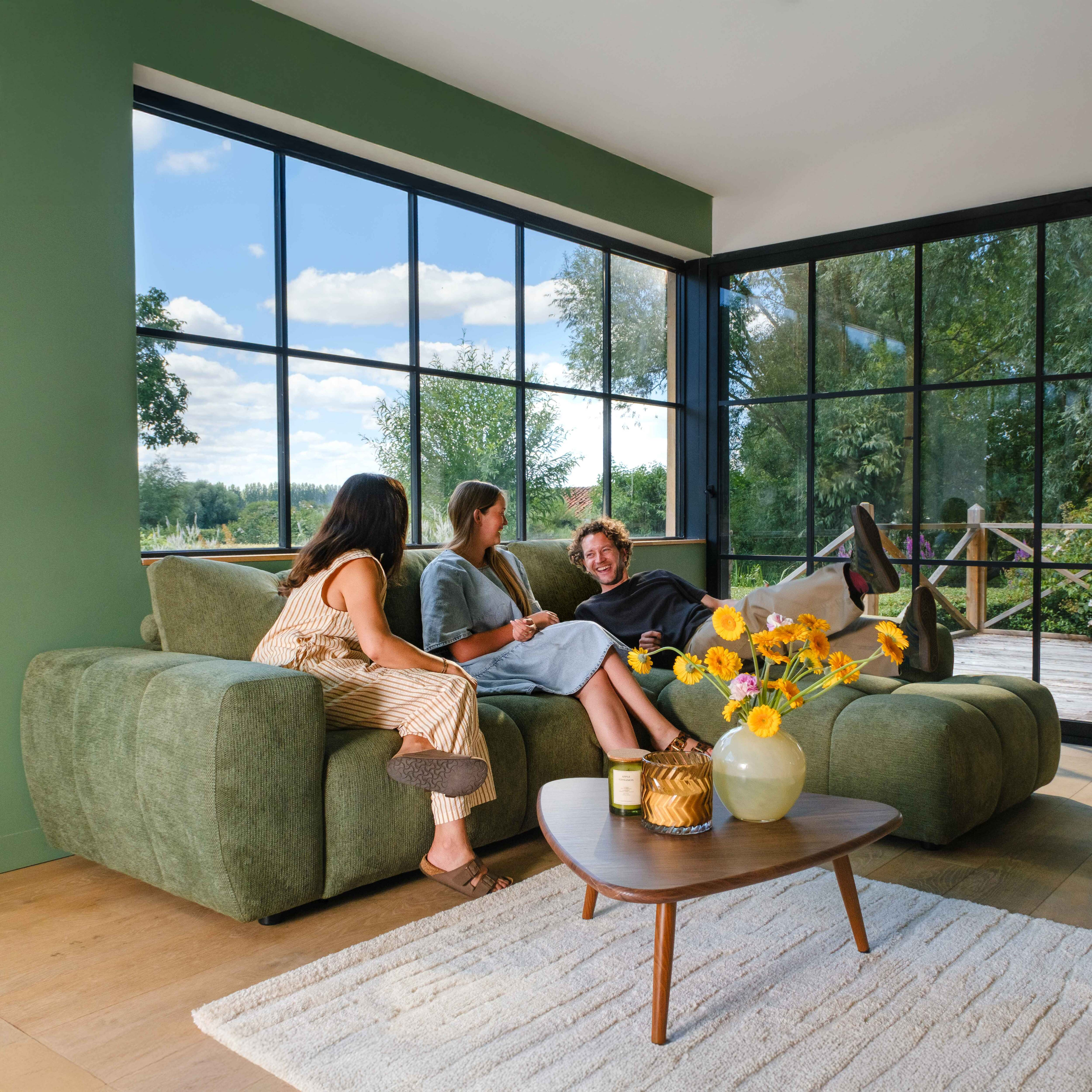 Drei Freunde entspannen auf einem grünen Sofa in einem sonnigen Raum mit großen Fenstern und Gartenblick.