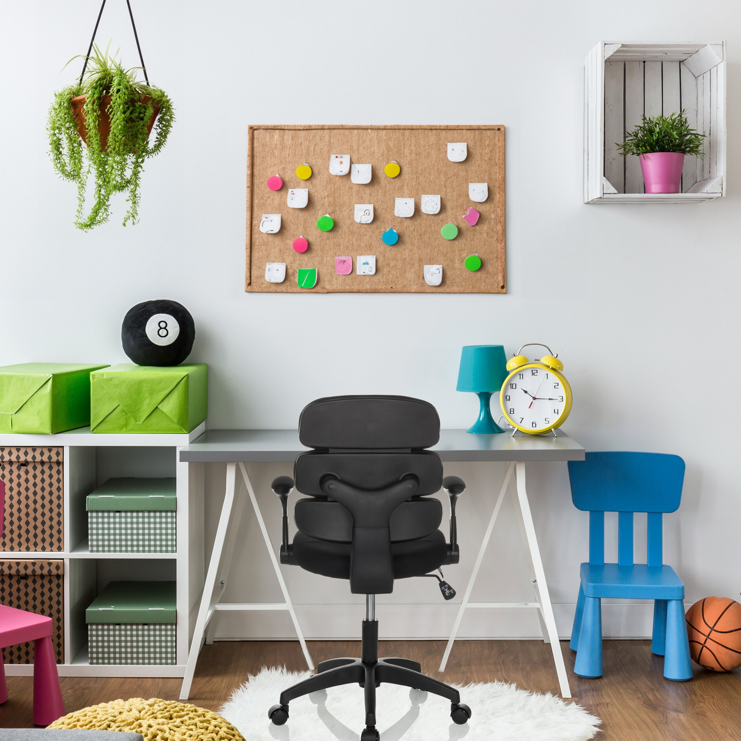 Ein Kinderschreibtisch mit schwarzem Bürostuhl, Korkwand, blauem Stuhl, Lampe und Uhren vor weißer Wand.