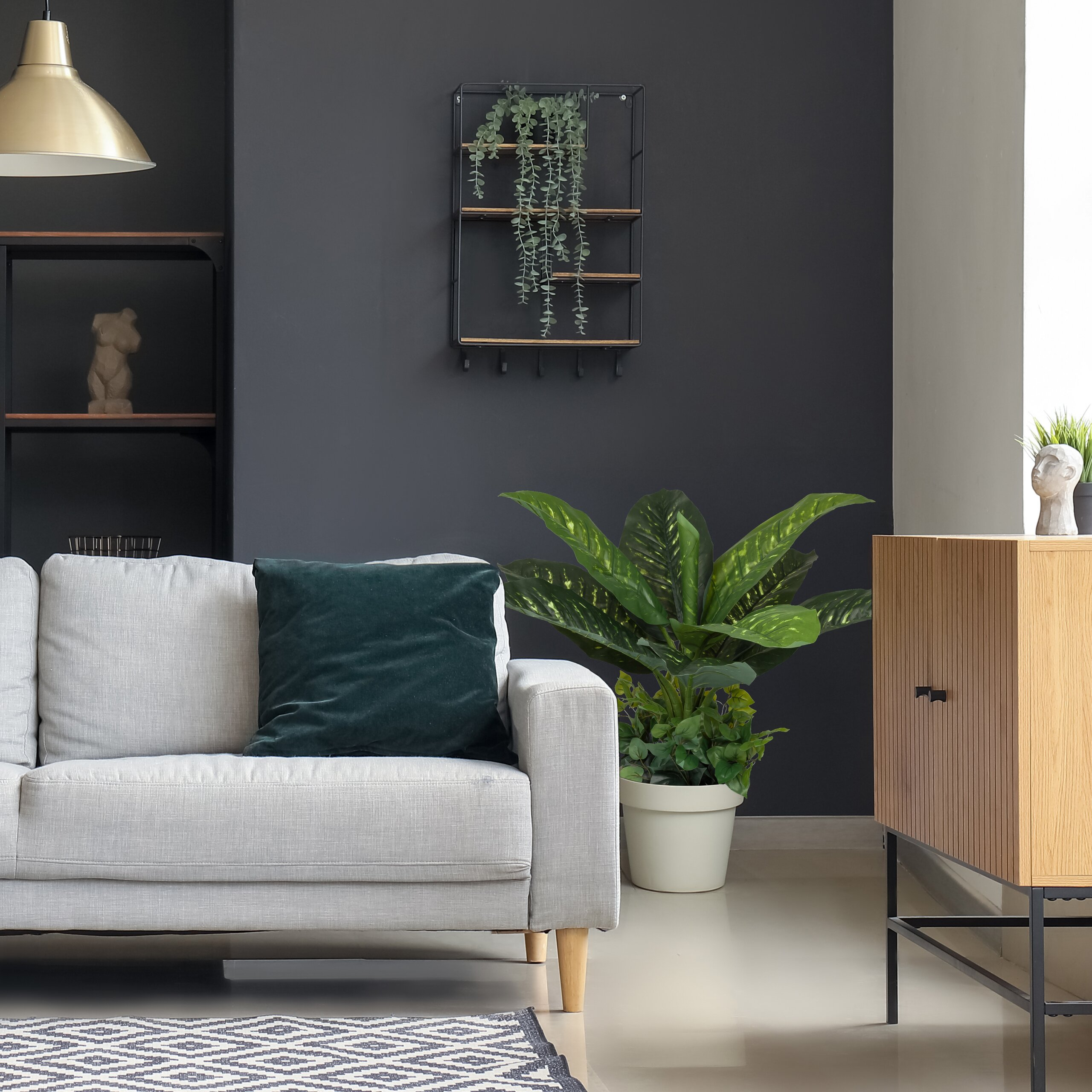 Ein graues Sofa mit Kissen, Pflanzen in Töpfen, einem Regal an der Wand und einem Holzsuschrank vor dunkler Wand.
