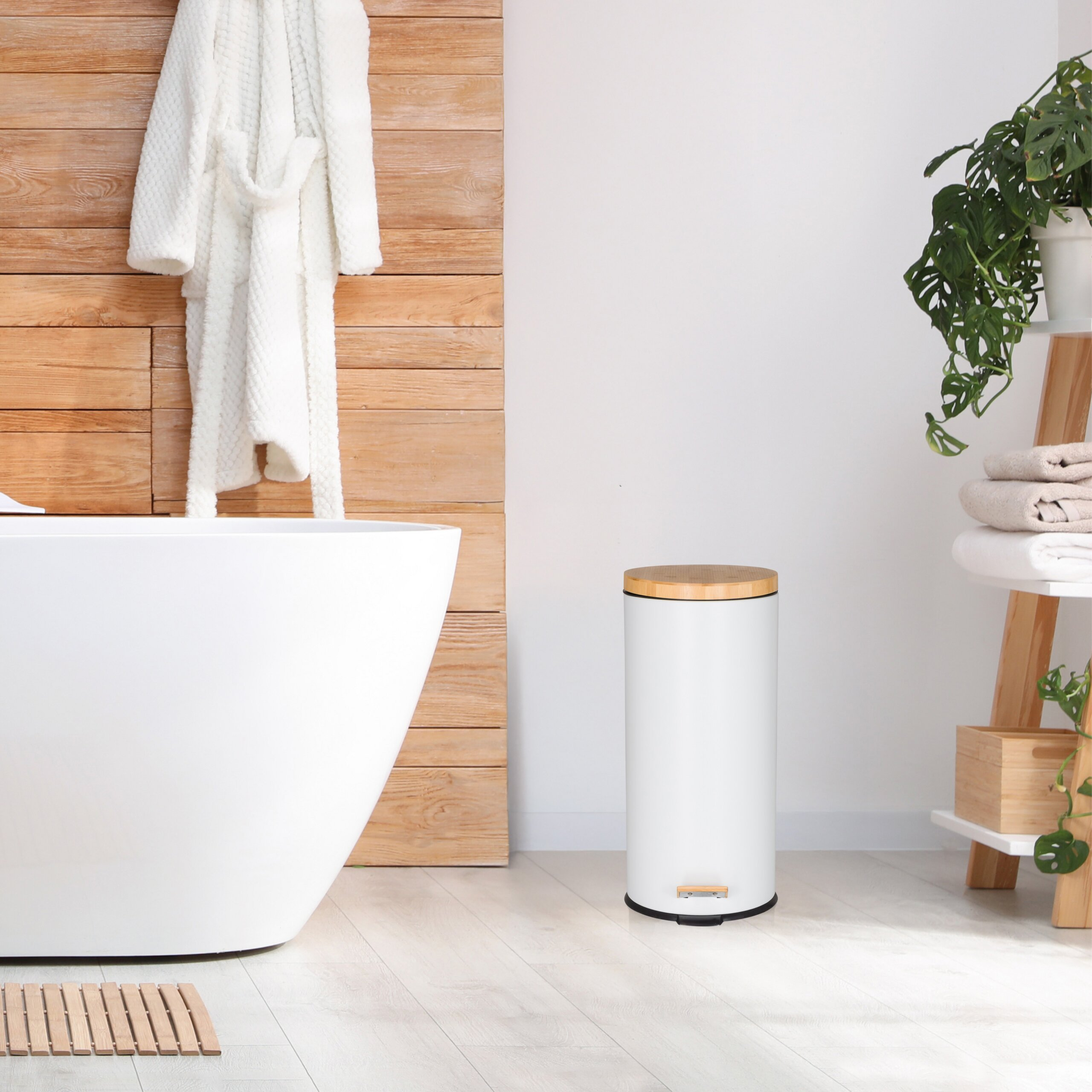 Eine weiße Badewanne, Holzwand mit weißem Bademantel, ein weißer Mülleimer mit Holzdeckel und Handtuchregal.