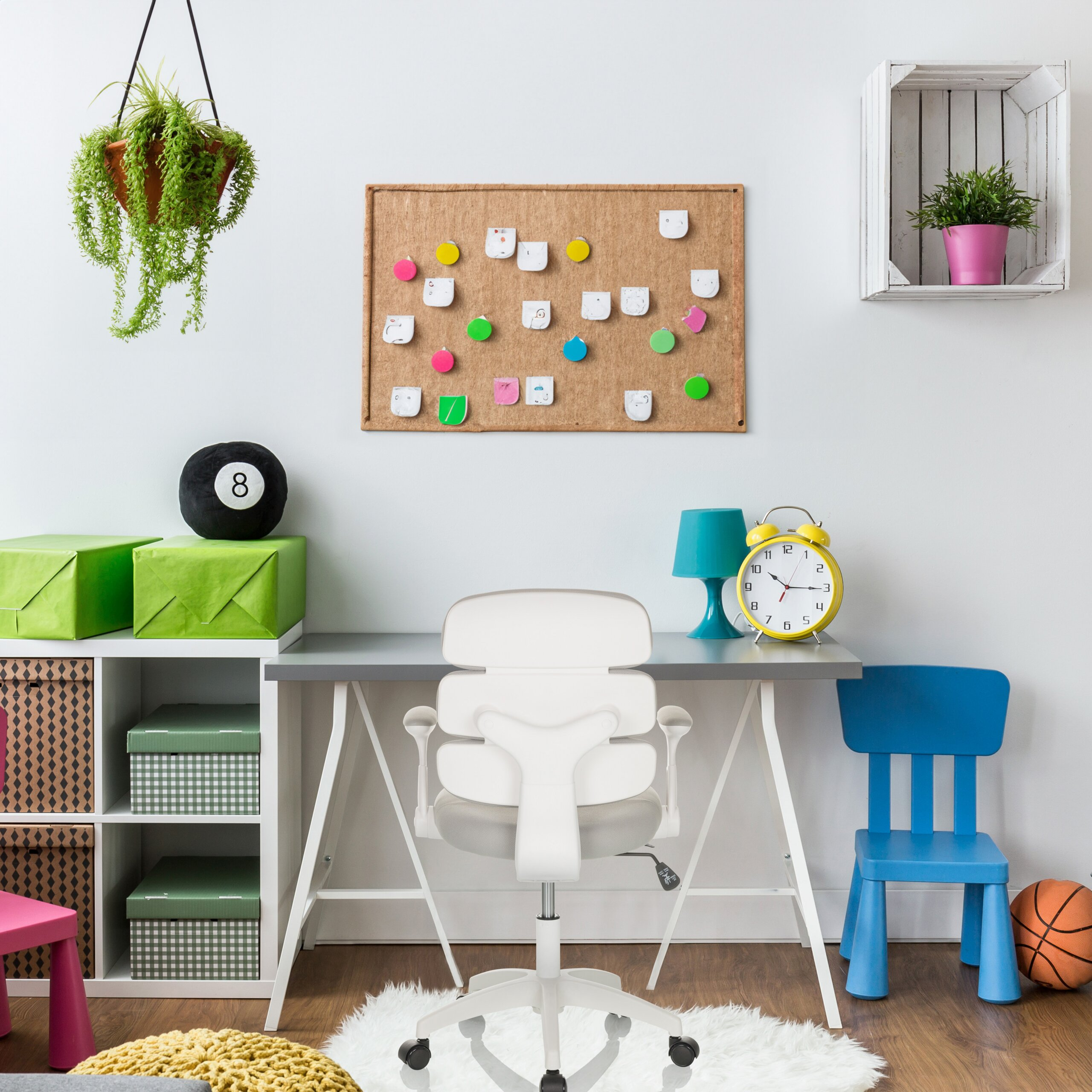 Ein helles Kinderzimmer mit weißem Schreibtisch, weißem Stuhl, bunten Kisten, Korktafel, Lampe und Pflanzen.
