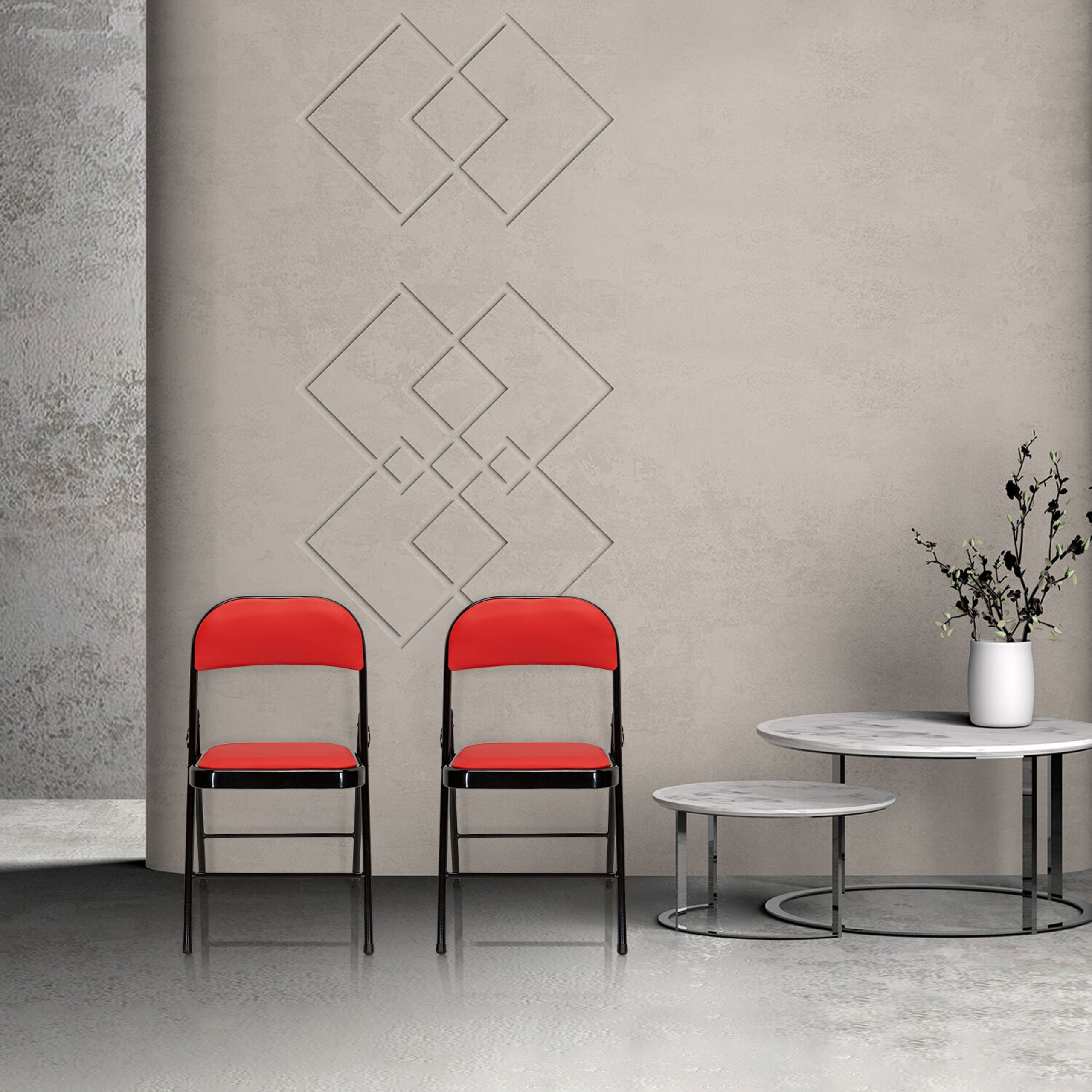 Zwei rote Stühle und zwei runde Tische stehen vor einer grauen Wand mit geometrischem Relief.