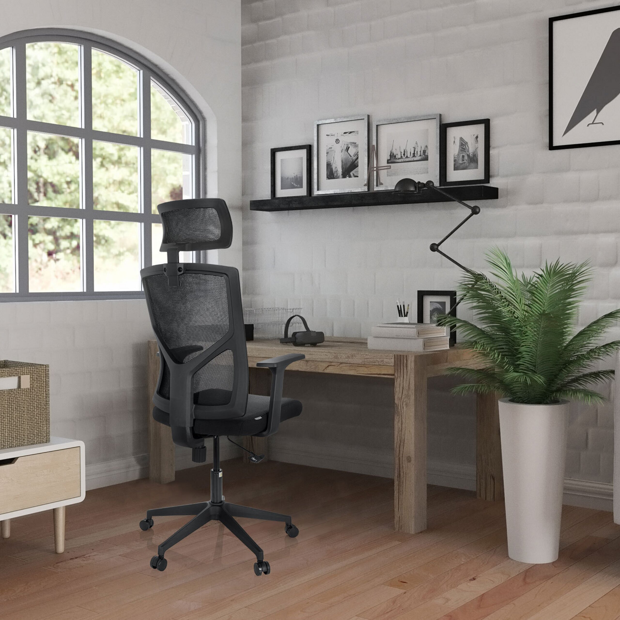 Ein schwarzer Bürostuhl steht vor einem Holztisch mit Pflanzen, Büchern und eingerahmten Bildern an der weißen Backsteinwand.