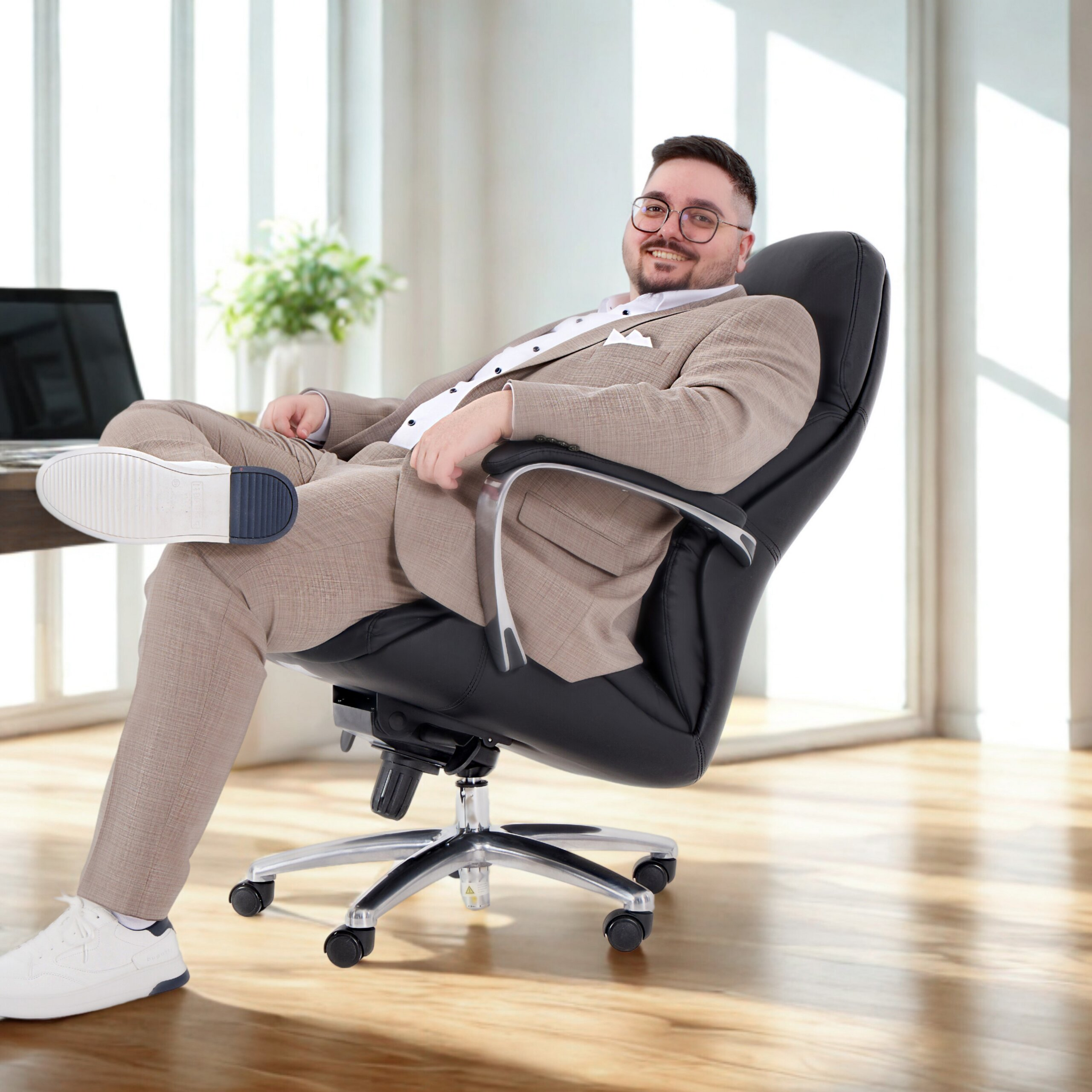 Ein lächelnder Mann in einem beigen Anzug und weißen Sneakers sitzt entspannt in einem schwarzen Chefsessel im Büro.