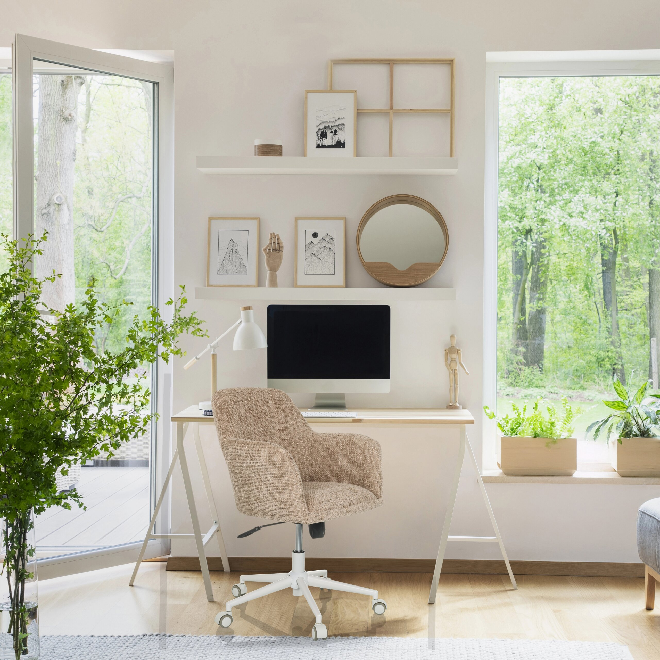Ein helles Heimbüro mit Computertisch, beige Sessel, Regalen und großen Fenstern mit Blick auf grüne Bäume.
