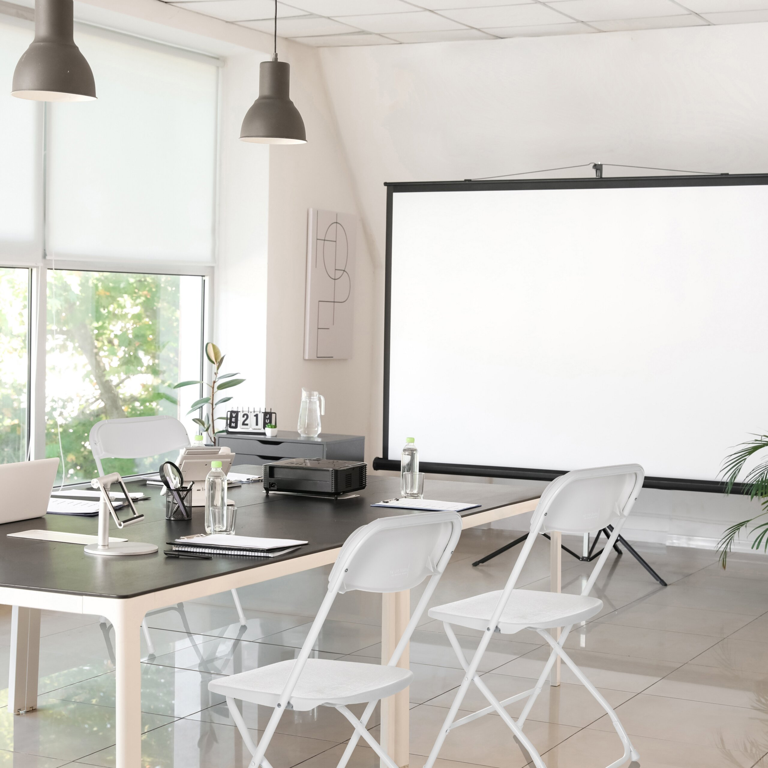 Heller Konferenzraum mit schwarzem Tisch, weißen Stühlen, Leinwand und großen Fenstern mit Grünblick.