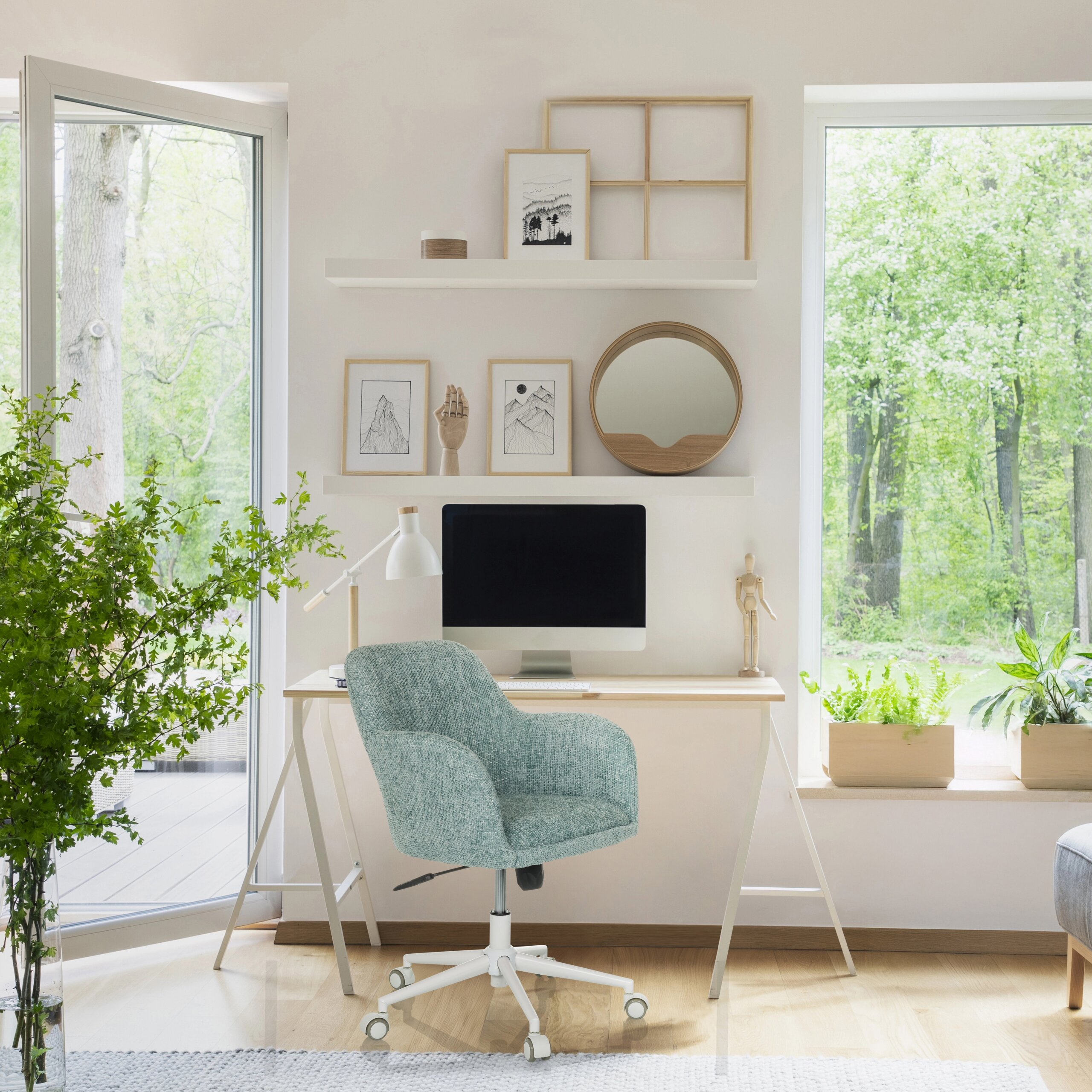 Ein heller Arbeitsplatz mit Schreibtisch, mintfarbenem Stuhl, Computer und Regalen vor großen Fenstern mit Waldblick.