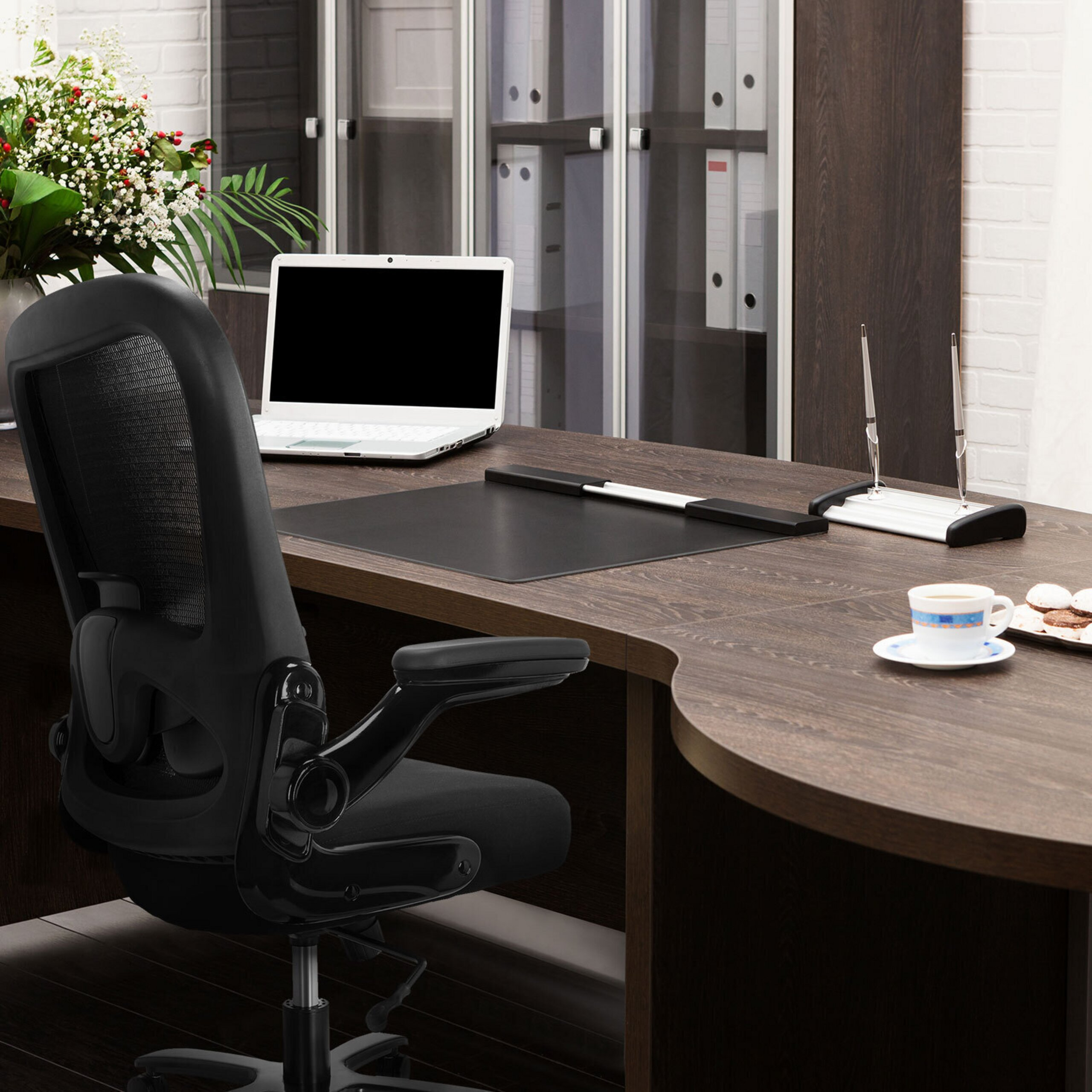 Moderne Bueroumbgebung mit einem schwarzen Buerostuhl, einem Laptop auf einem Holztisch und Blumen im Hintergrund.